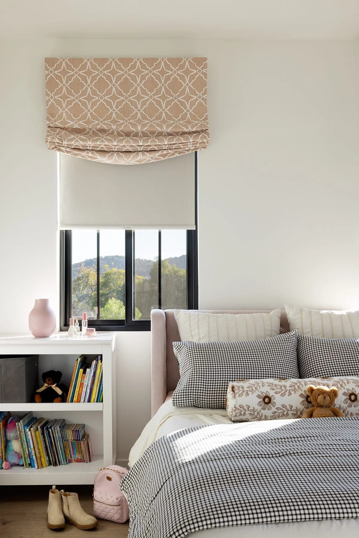 A bedroom with a window showing trees and hills outside. The bed has checkered and floral pillows, a teddy bear, and a cream-colored quilt. Next to the bed is a white bookshelf with books and decorative items, a pink vase, and a small pink backpack on the floor. A pair of beige boots is beside the backpack.