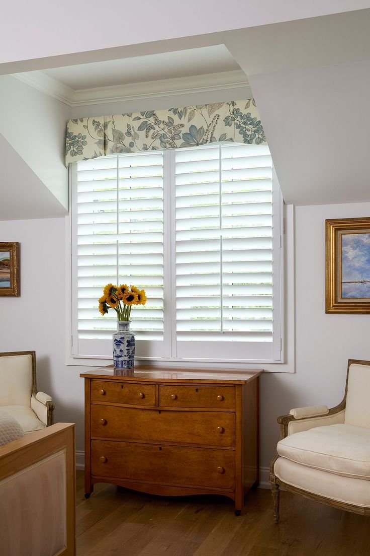 Living room corner with a large window having white plantation shutters, floral valance, wooden dresser with sunflower bouquet in a blue-and-white vase, vintage armchair and framed landscape paintings.