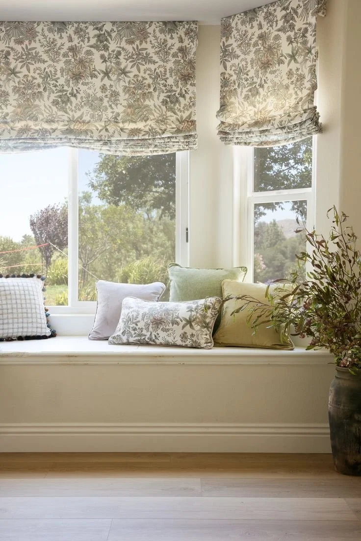 A window seat with floral-patterned curtains and several decorative pillows, overlooking a garden with trees and greenery, with a large potted plant nearby.