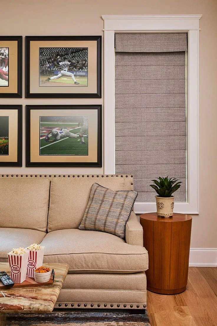 A beige sofa with a plaid cushion, next to a round wooden side table with a potted plant, topped with framed sports photographs on the wall, popcorn and snacks on a marble coffee table, and a window with a gray roller shade.