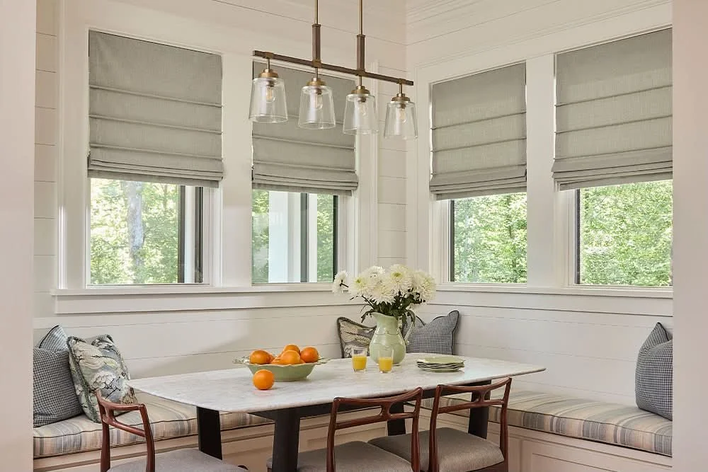 A cozy kitchen nook with white paneled walls, a marble table, gray window shades, three windows, a flower arrangement, and seating with pillows.