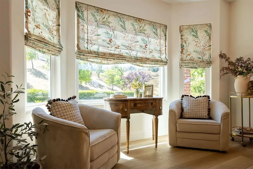 A cozy living room corner with two white armchairs with checkered pillows, a wooden side table with plants and a picture frame, a gold cart with a large vase and flowers, and windows with floral Roman shades letting in natural light.