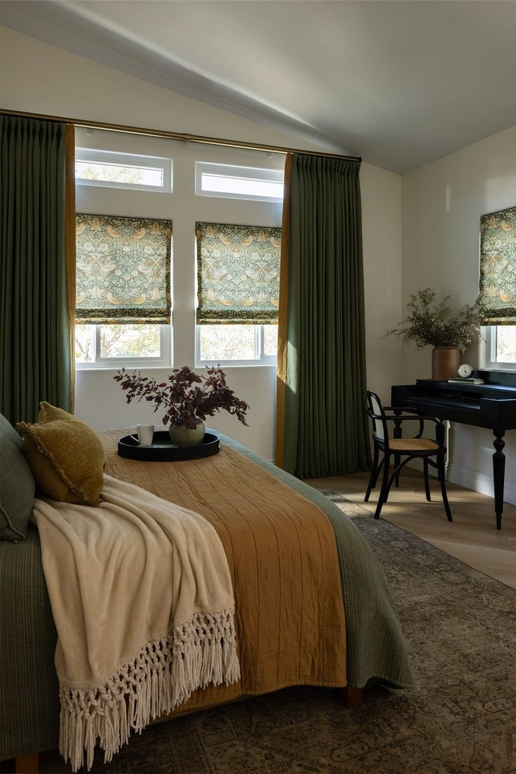 Cozy bedroom with green curtains, patterned roman shades, a bed with mustard and cream bedding, a black writing desk with a chair, and decorative plants.