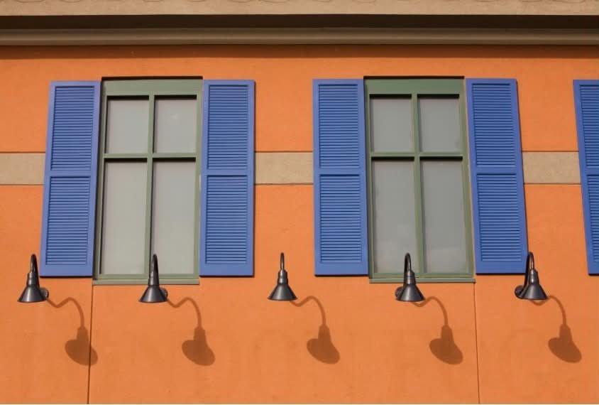 Orange building wall with three windows having blue shutters, each with a black lampshade attached below, casting shadows on the wall.