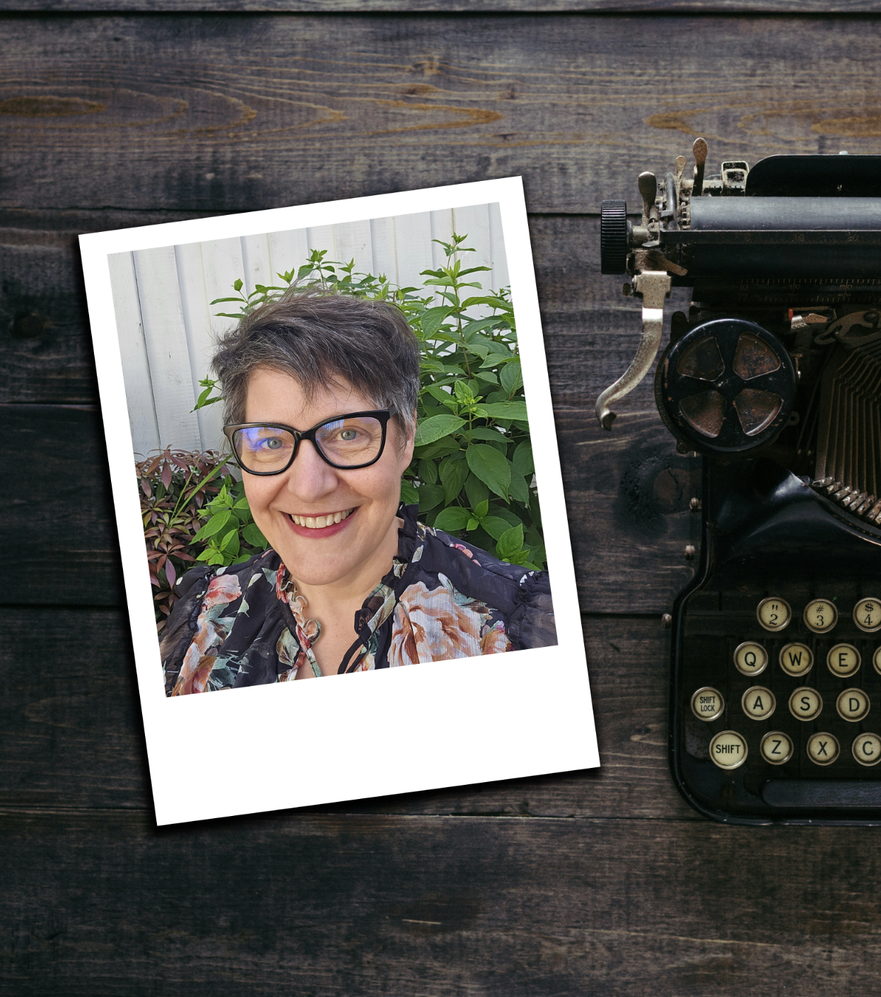 Vintage typewriter next to a printed photo of a smiling woman with short dark hair and glasses, standing in front of greenery and a white fence, on a dark wooden surface.