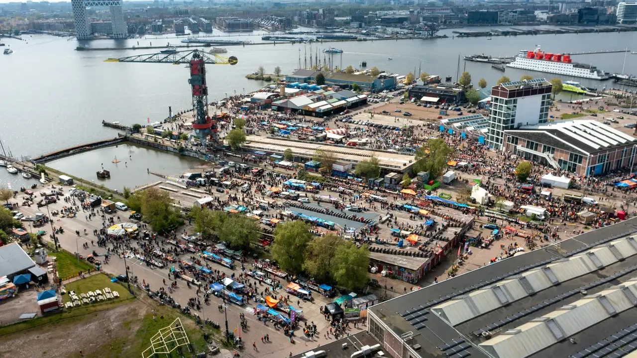 Overview of a NDSM wharf, busy market square along a river with many people, market stalls, a large tower with crane, boats on the river and a modern facade with many windows.