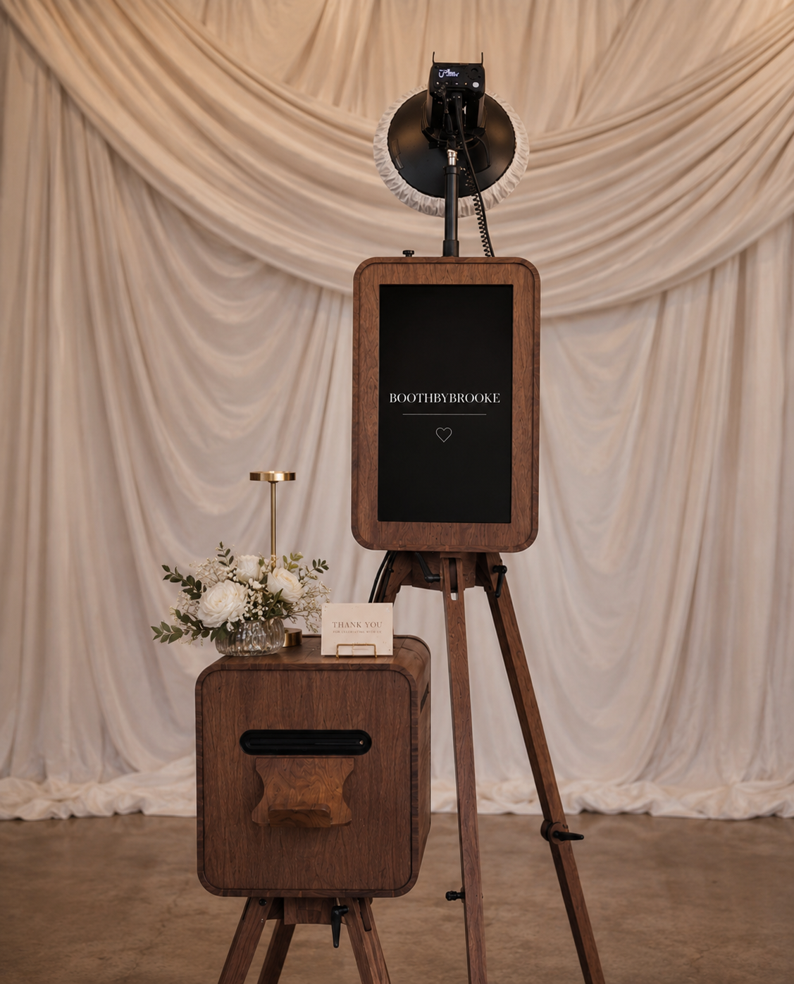 Photo booth setup with a large camera and a wooden stand, a smaller wooden table with flowers and a thank you card, and a backdrop of elegant cream-colored drapes.
