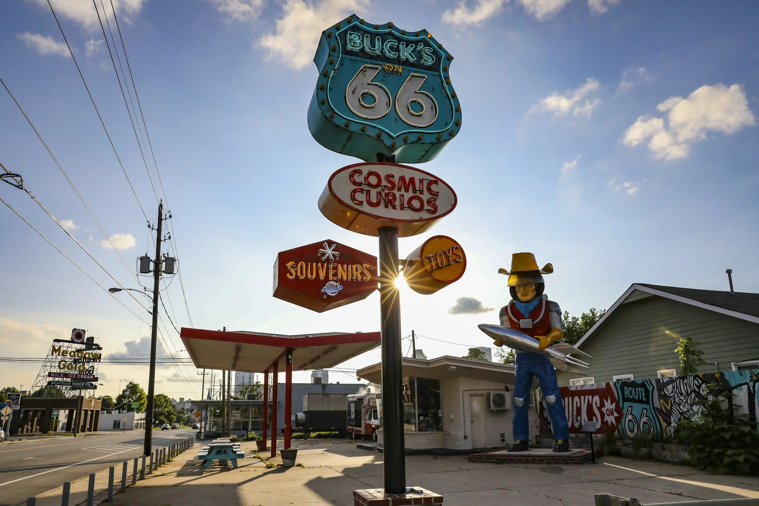 A vintage Route 66 sign with the words "Buck's on 66" illuminated at the top, and a smaller sign underneath that reads "Cosmic Curios". There is a statue of a cowboy with a hat, holding a surfboard, standing beside the sign. The background shows a small building, a street, power lines, and a partly cloudy sky during sunset.