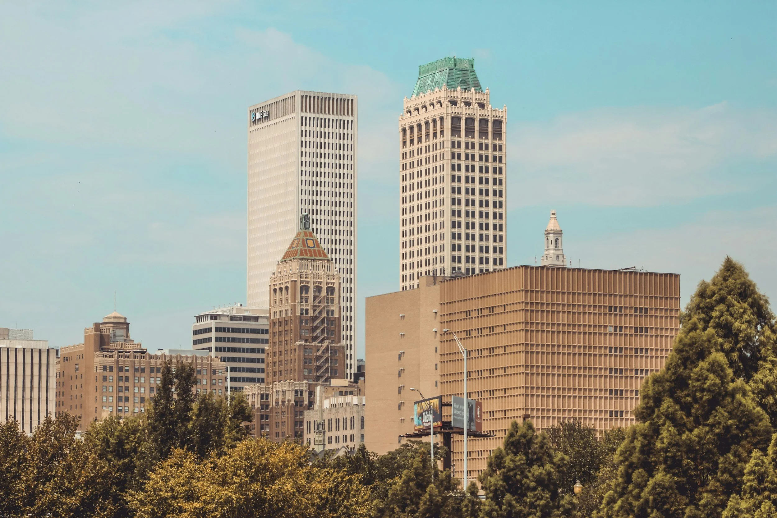A city skyline with tall skyscrapers, including two prominent buildings with unique architectural styles, and trees in the foreground under a partly cloudy sky.