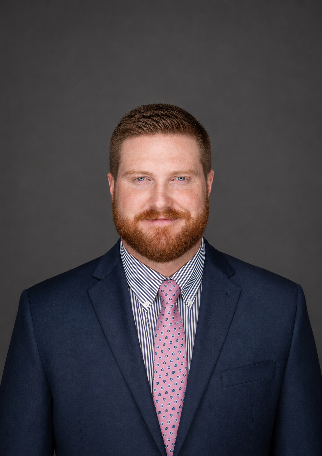 Headshot of a man with red hair and beard wearing a navy suit, striped shirt, and pink tie, against a dark gray background.