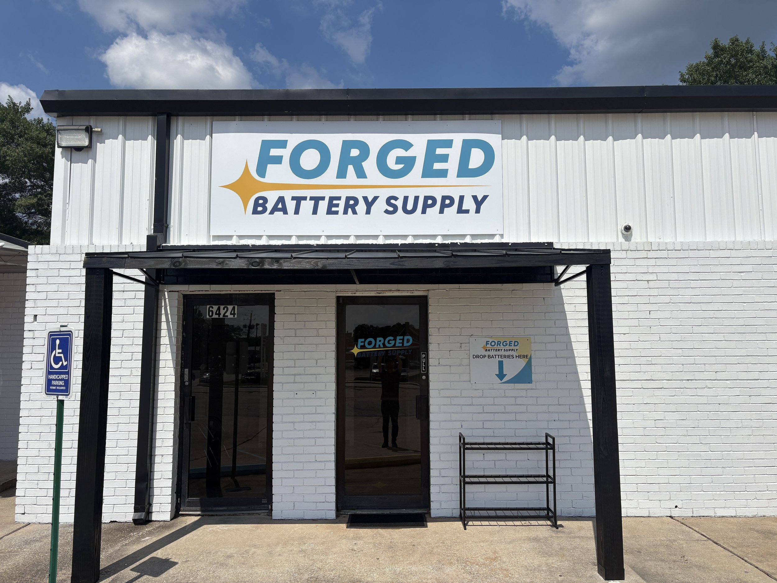 Storefront with sign reading 'Forged Battery Supply' and a blue handicapped parking sign to the left.
