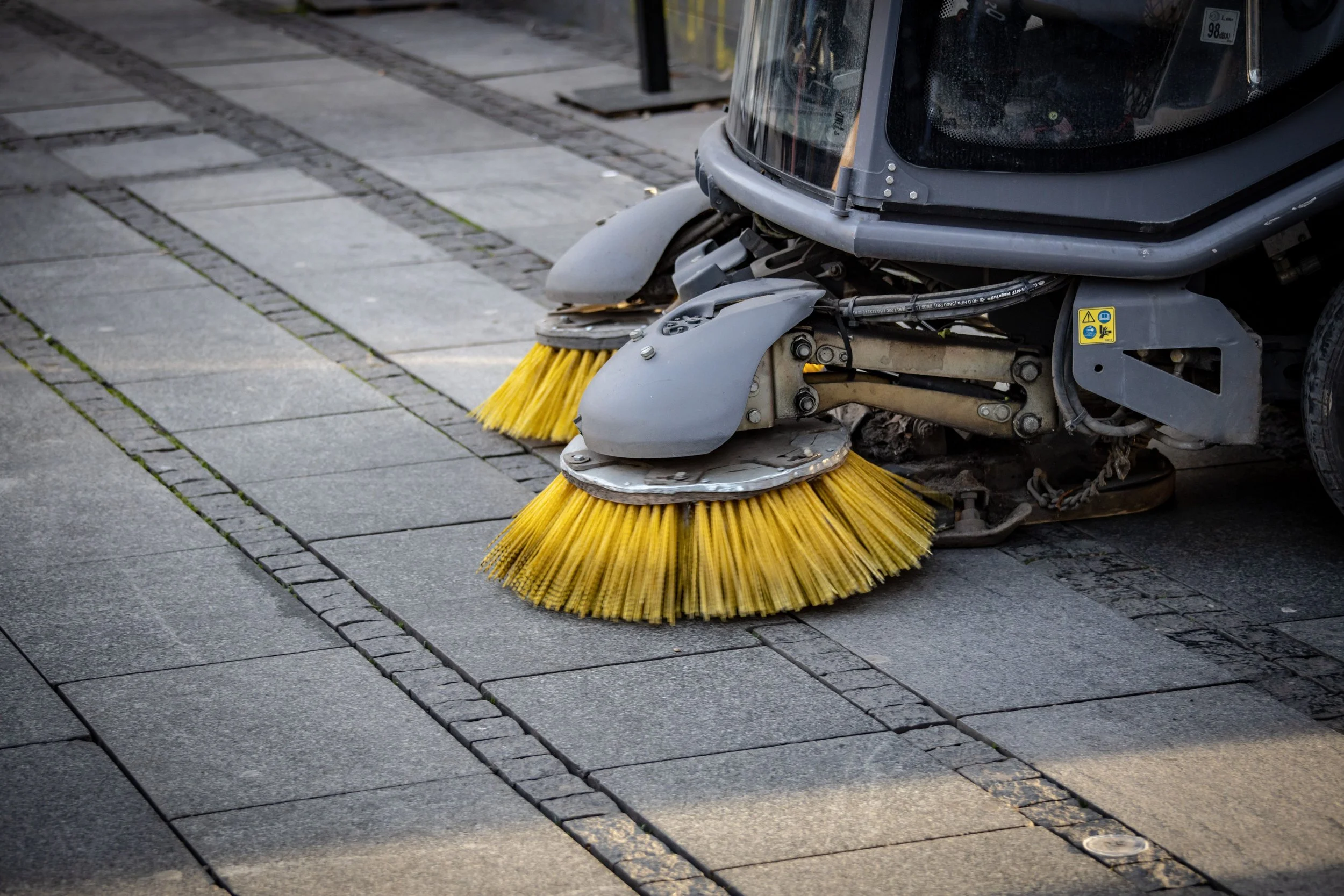 A street sweeper machine clearing debris from the gray brick sidewalk.