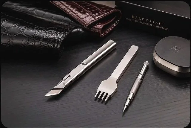 A collection of luxury accessories and tools on a black surface, including two leather wallets, a box with the label 'Built to Last', a fountain pen, a fork, and an electric shaver.