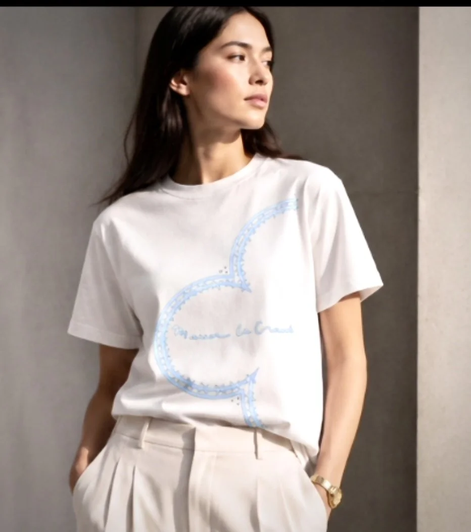 A woman with long dark hair wearing a white T-shirt with a blue design and beige pants, standing indoors with one hand in her pocket and looking to the side.