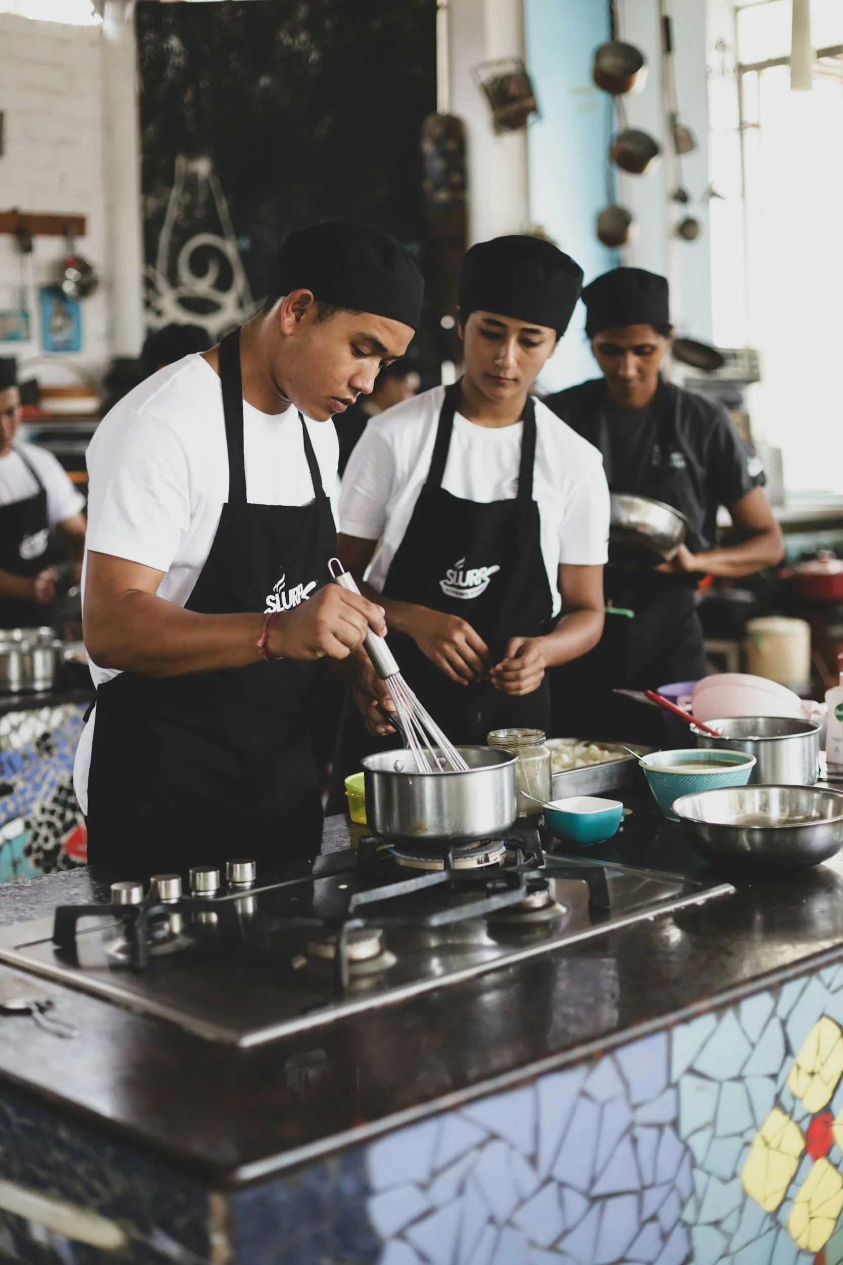 Three young chefs in black hats and white aprons preparing food in a modern kitchen with cooking utensils and ingredients on a stove.