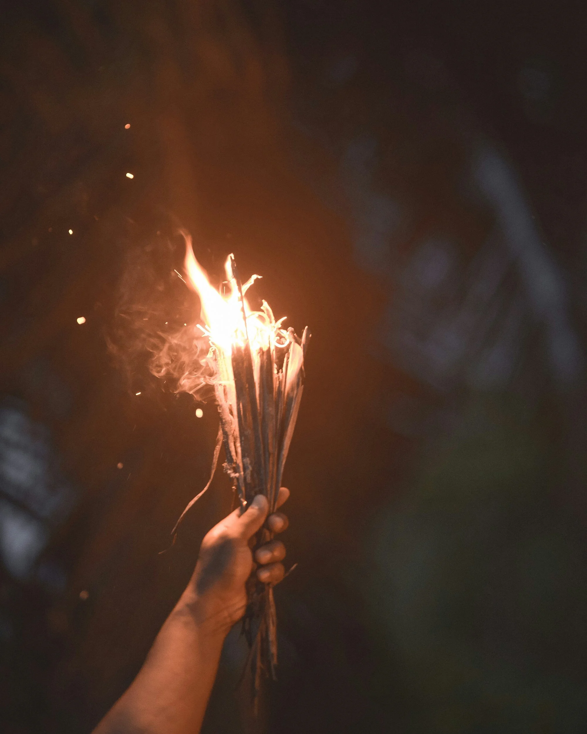 A person's hand holding a lit sparkler firework during nighttime, with sparks flying upwards against a dark sky.
