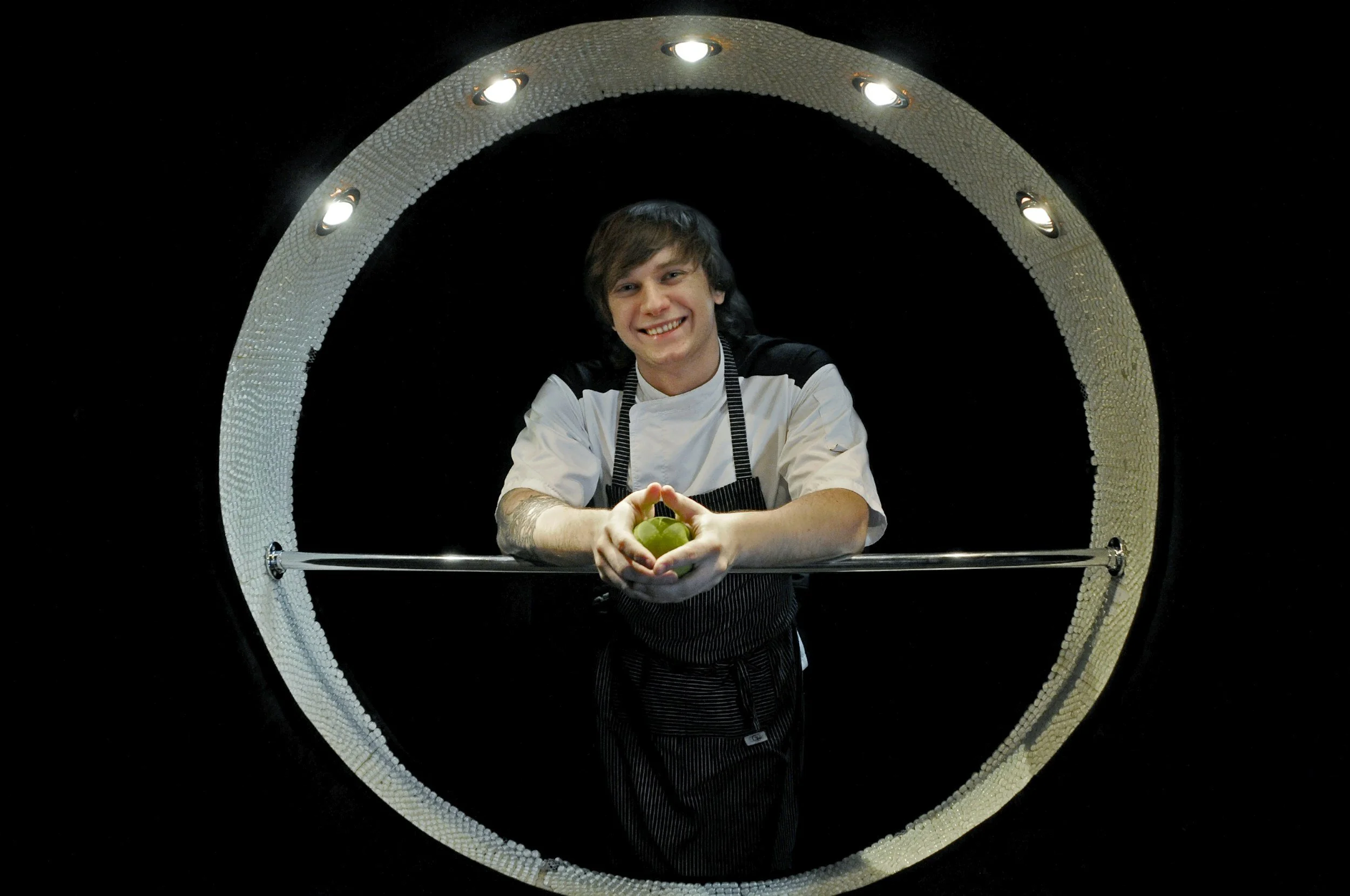 A young man with a tattoo on his right arm, smiling and holding a green apple with both hands, standing behind a circular opening in a reflective surface, wearing a white shirt and black apron. The background is black with the circular opening framed by a textured, metallic ring with built-in lights.