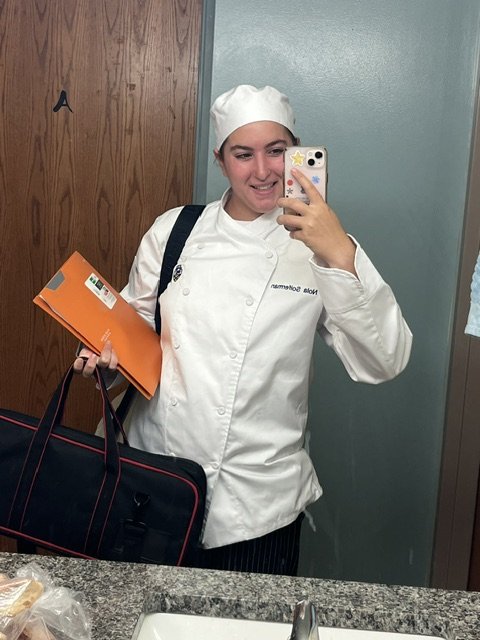 Young woman in a chef's uniform and hat taking a mirror selfie in a bathroom.