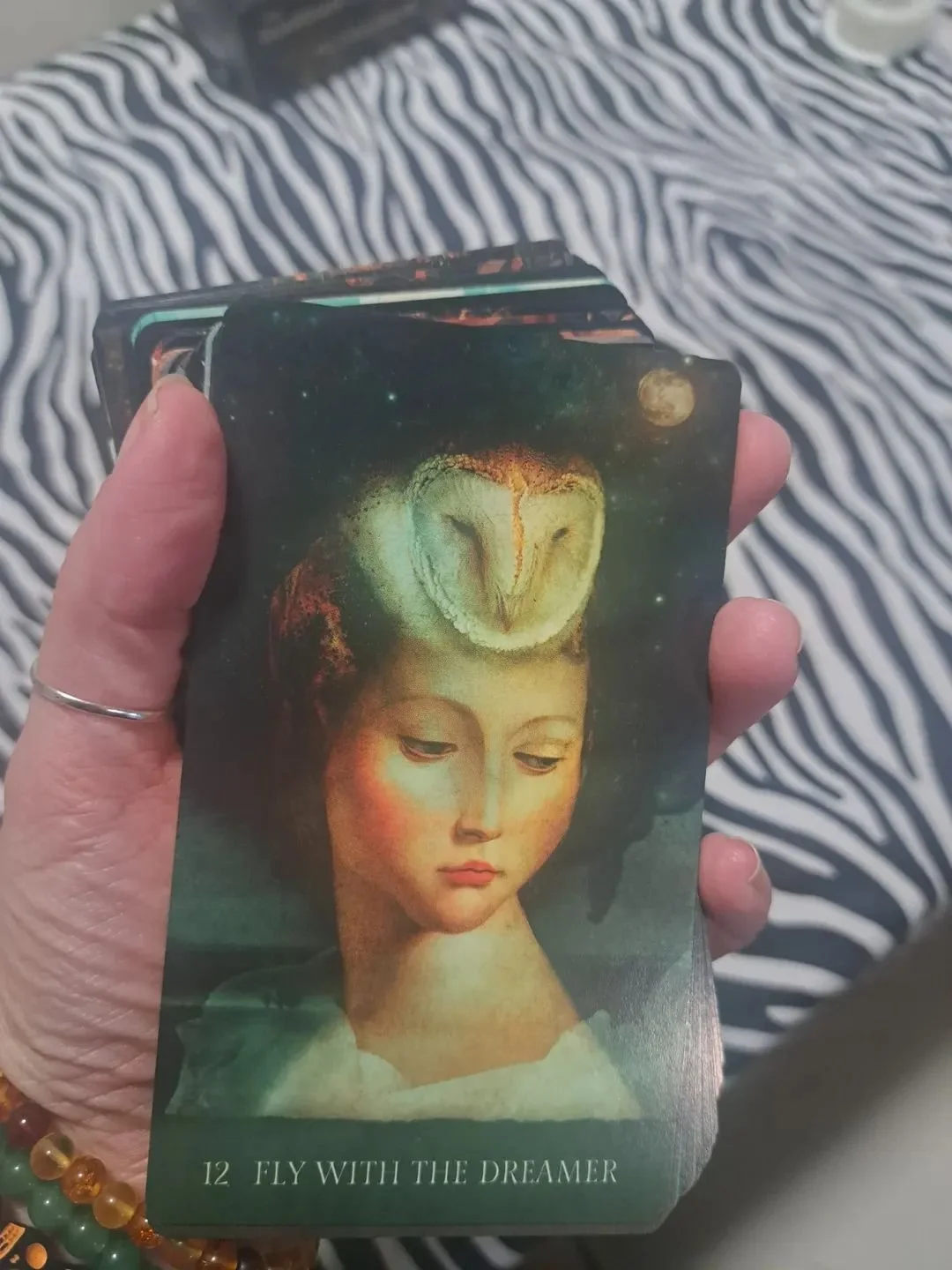 A person holding a deck of oracle cards with mystical art, featuring an angelic figure and an owl, titled 'Fly with the Dreamer', number 12.