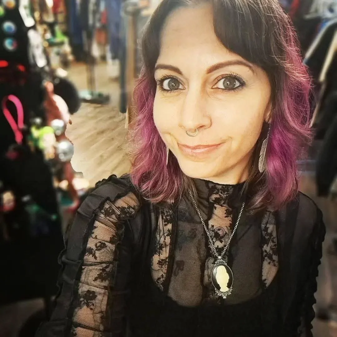 A woman with pink and brown hair, wearing a black lace top and a necklace with a cameo pendant, smiling in a store.