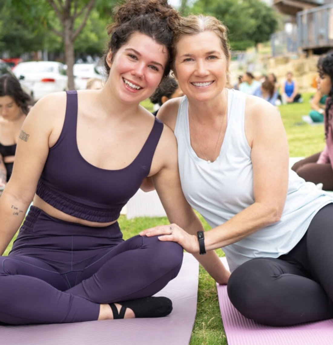 Two women smiling, sitting on yoga mats outdoors during a yoga class, surrounded by other participants practicing yoga.