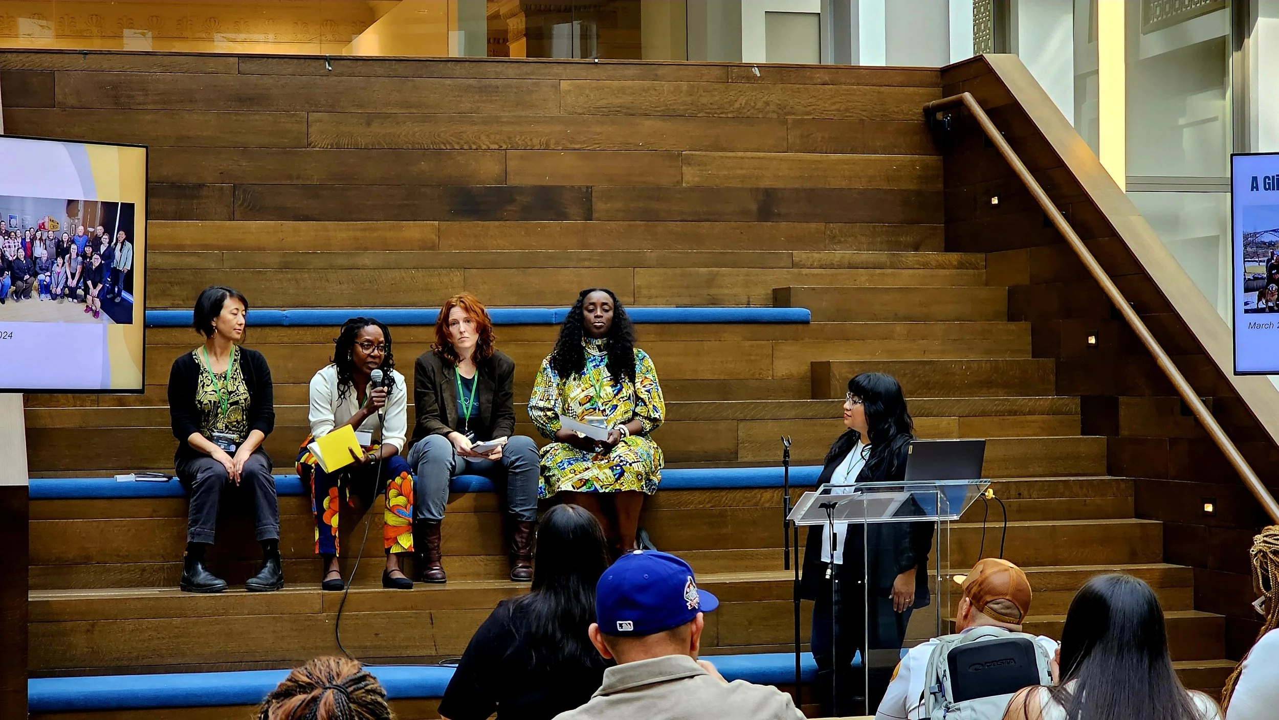 A panel of five women seated on a blue bench with wooden steps behind them, participating in a discussion at an indoor event. There are two large screens displaying images and text to their sides. The audience is visible in the foreground.