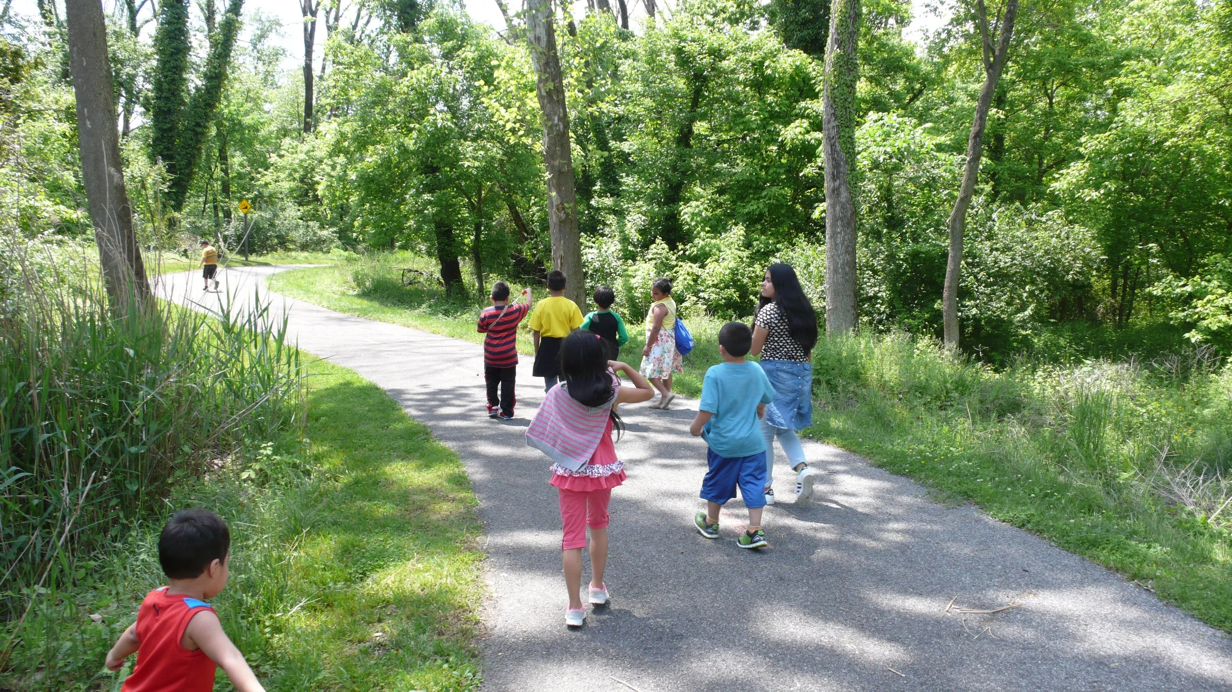 Children and a woman walking on a paved trail through a lush green forest in daytime.