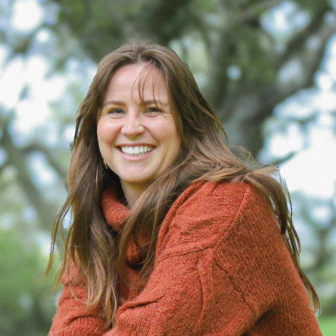 Sierra Mathias, Director of Movement Building and Advocacy, A smiling woman with long brown hair wearing a rust-colored sweater outdoors with a background of blurred trees.
