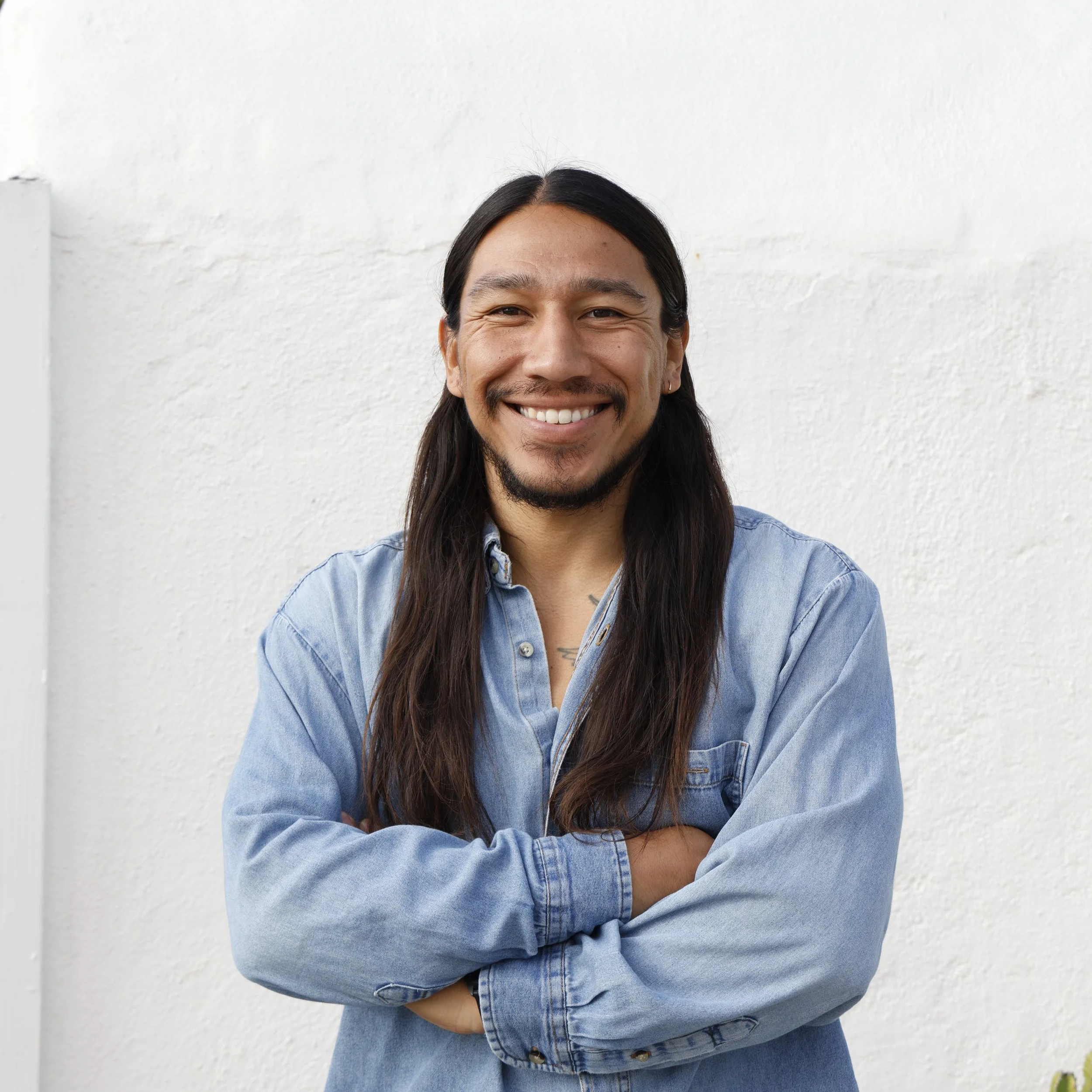Mario Ordoñez-Calderón, Executive Director  Un Mar de Colores, Close-up of a smiling person with long dark hair, wearing a light denim shirt, standing against a plain white wall.