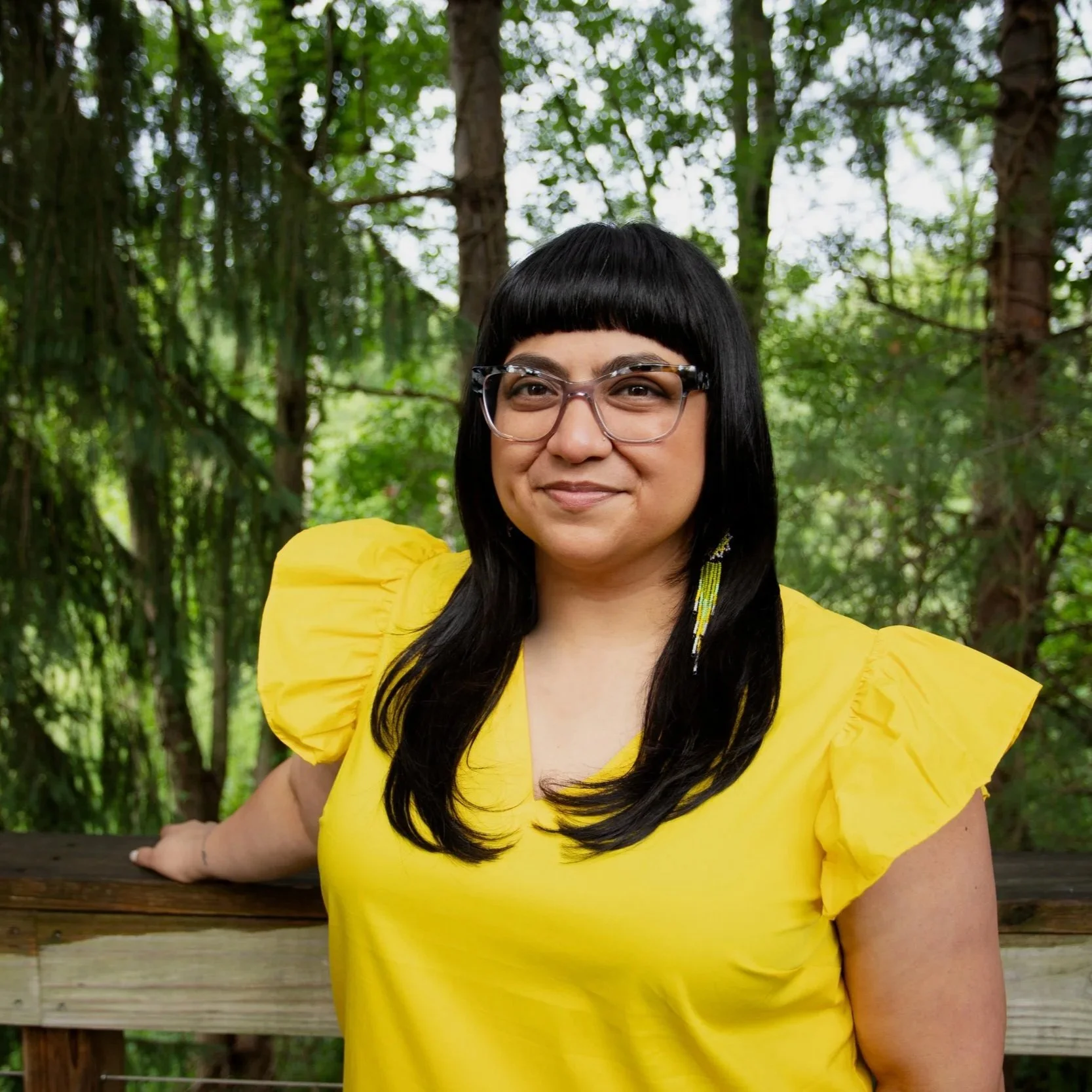 Betsy López-Wagner, Principal, Lopez-Wagner Strategies, A woman with black hair, glasses, and earrings, smiling, wearing a bright yellow dress with puffed sleeves, standing outdoors in a wooded area with trees and greenery.