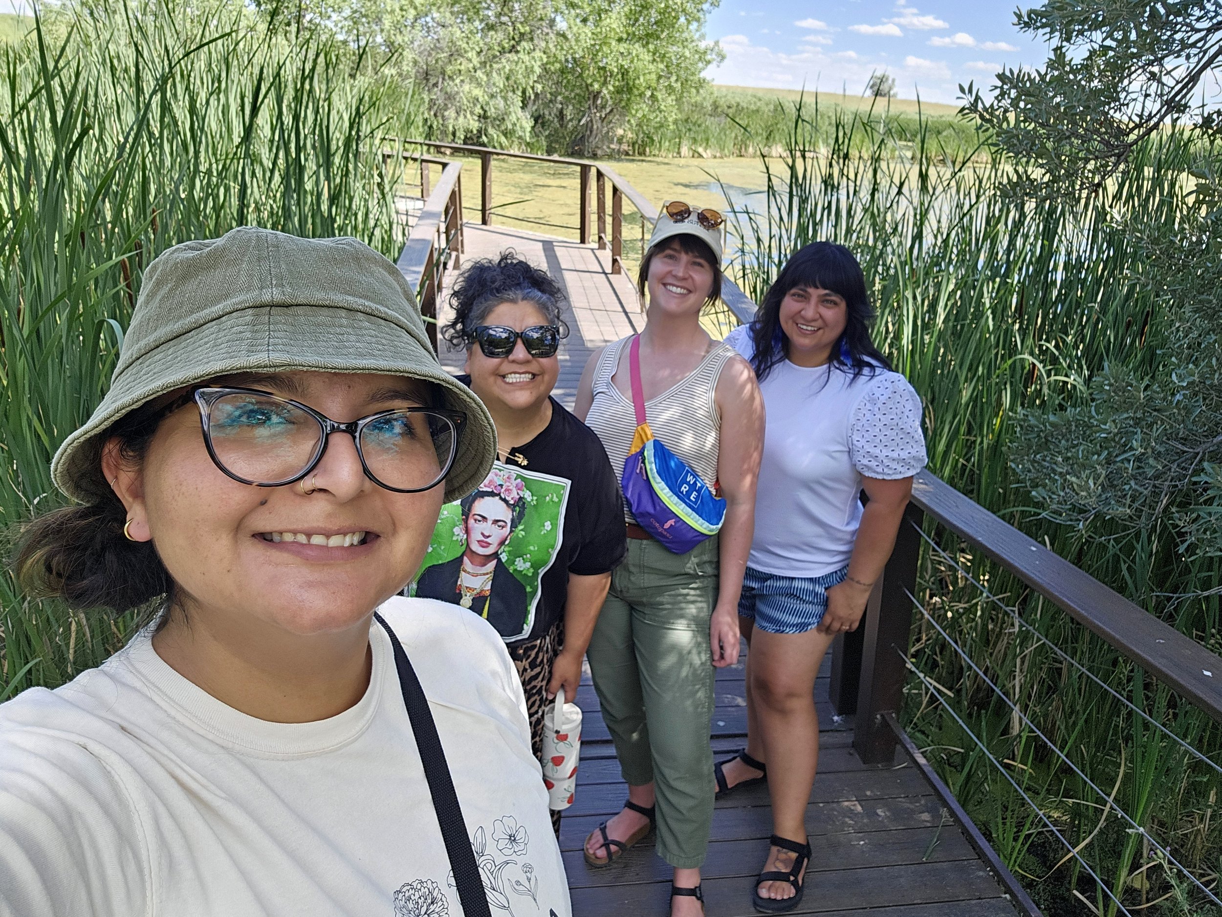 Group of four women standing on a wooden boardwalk surrounded by tall green reeds, near a pond or marsh, smiling and taking a selfie.