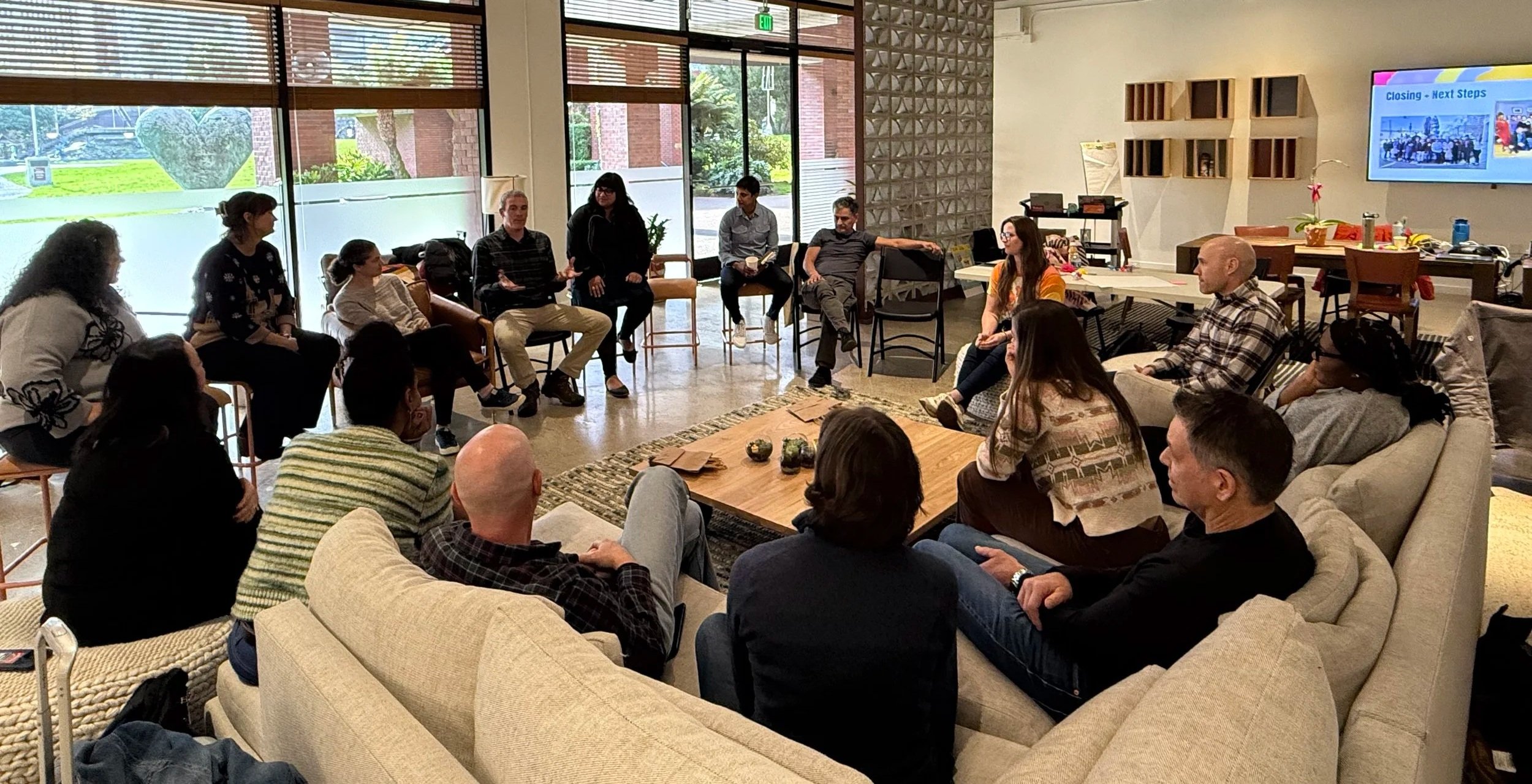 A group of diverse people sitting in a circle in a modern, well-lit meeting room with large windows and wooden blinds, engaging in a discussion or workshop.