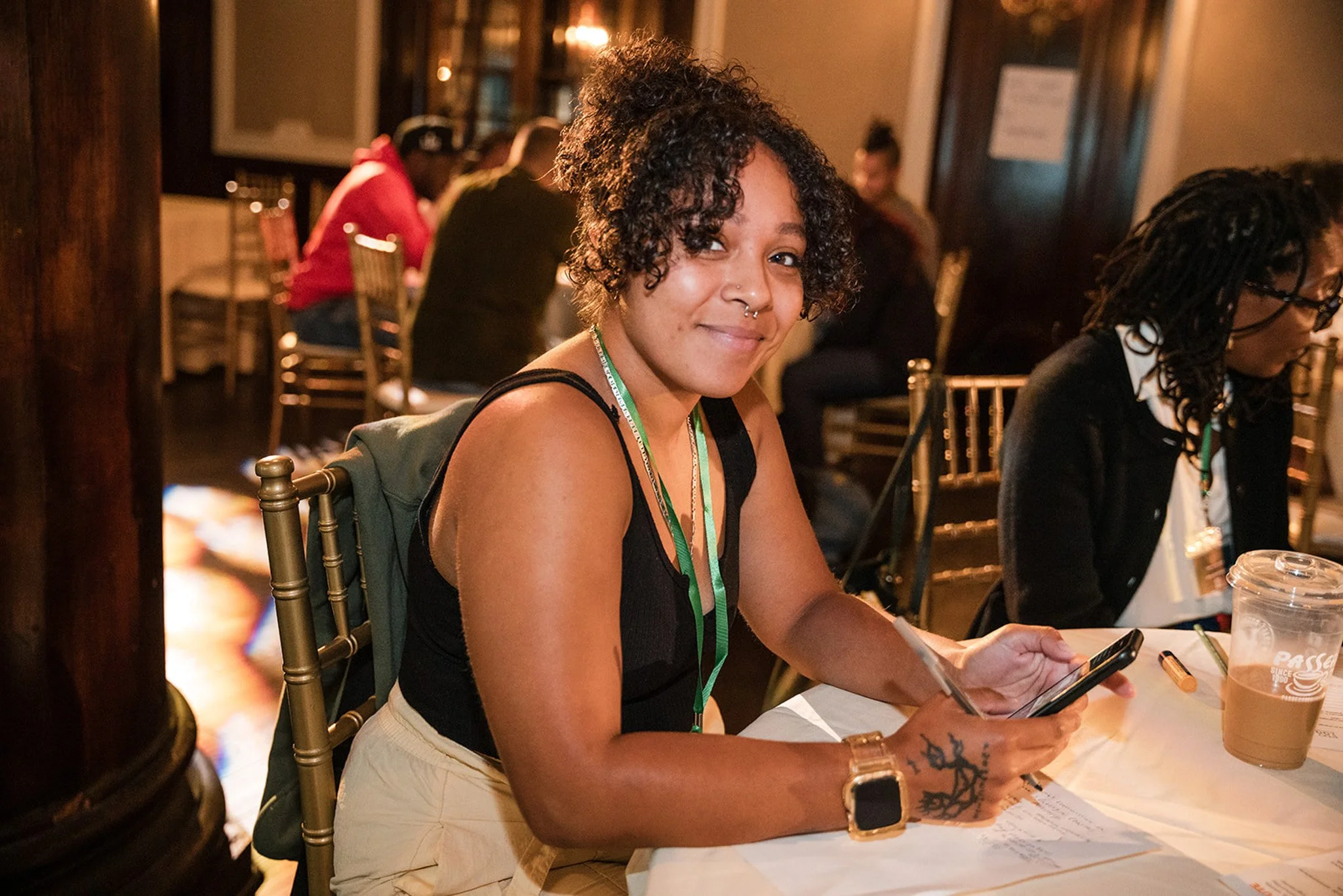 A young woman with curly hair and a septum piercing sitting at a table with a drink and a phone in her hand, smiling with other people in the background in a warmly lit indoor setting.