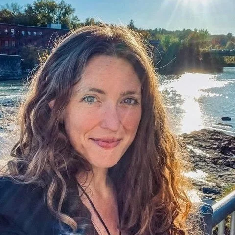 Kit Harrington, Director of Early Learning Policy  Natural Start Alliance, A woman with long curly brown hair smiling outdoors near a river with trees and buildings in the background, sunlight brightening the scene.