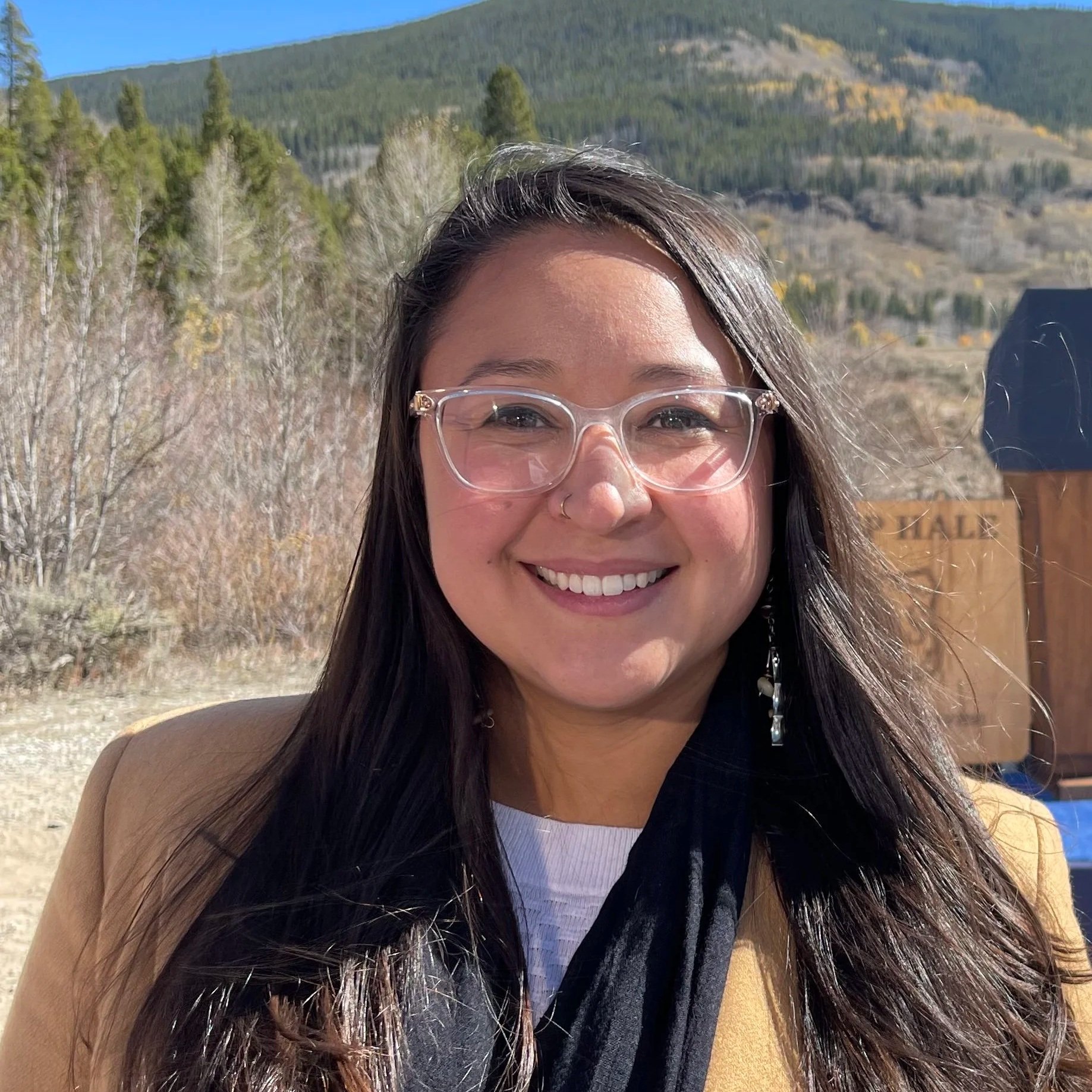 Chela Garcia Irlando, Executive Director  Next 100 Coalition, A woman smiling outdoors with mountain scenery, trees, and a wooden sign in the background.