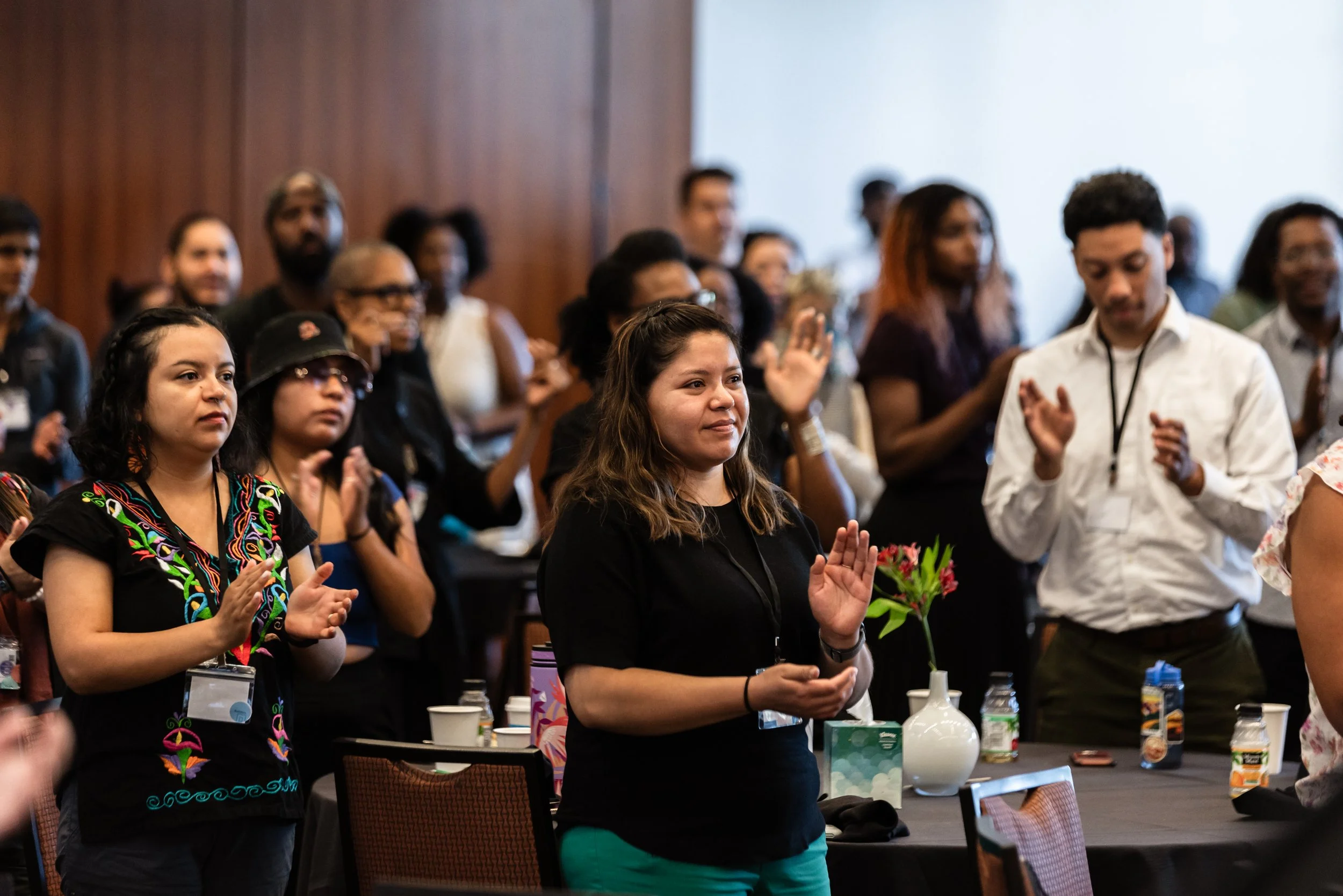 People attending an indoor event, standing and clapping, some with drinks and flowers on tables in front of them.