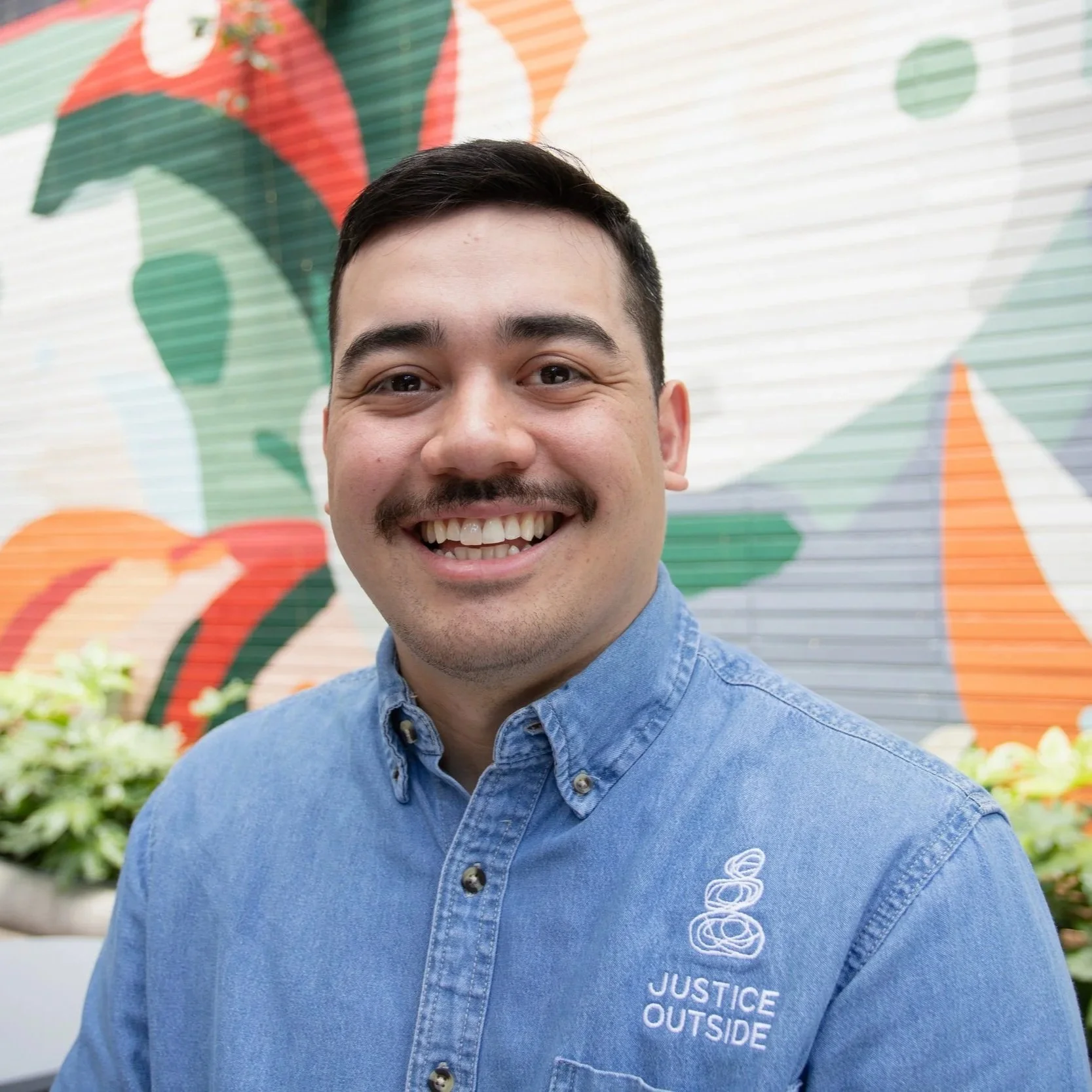 Ethan Metzger, Movement Building Coordinator, A smiling man with short dark hair and facial hair wearing a blue denim shirt standing in front of a colorful mural with green, orange, white, and red abstract shapes.