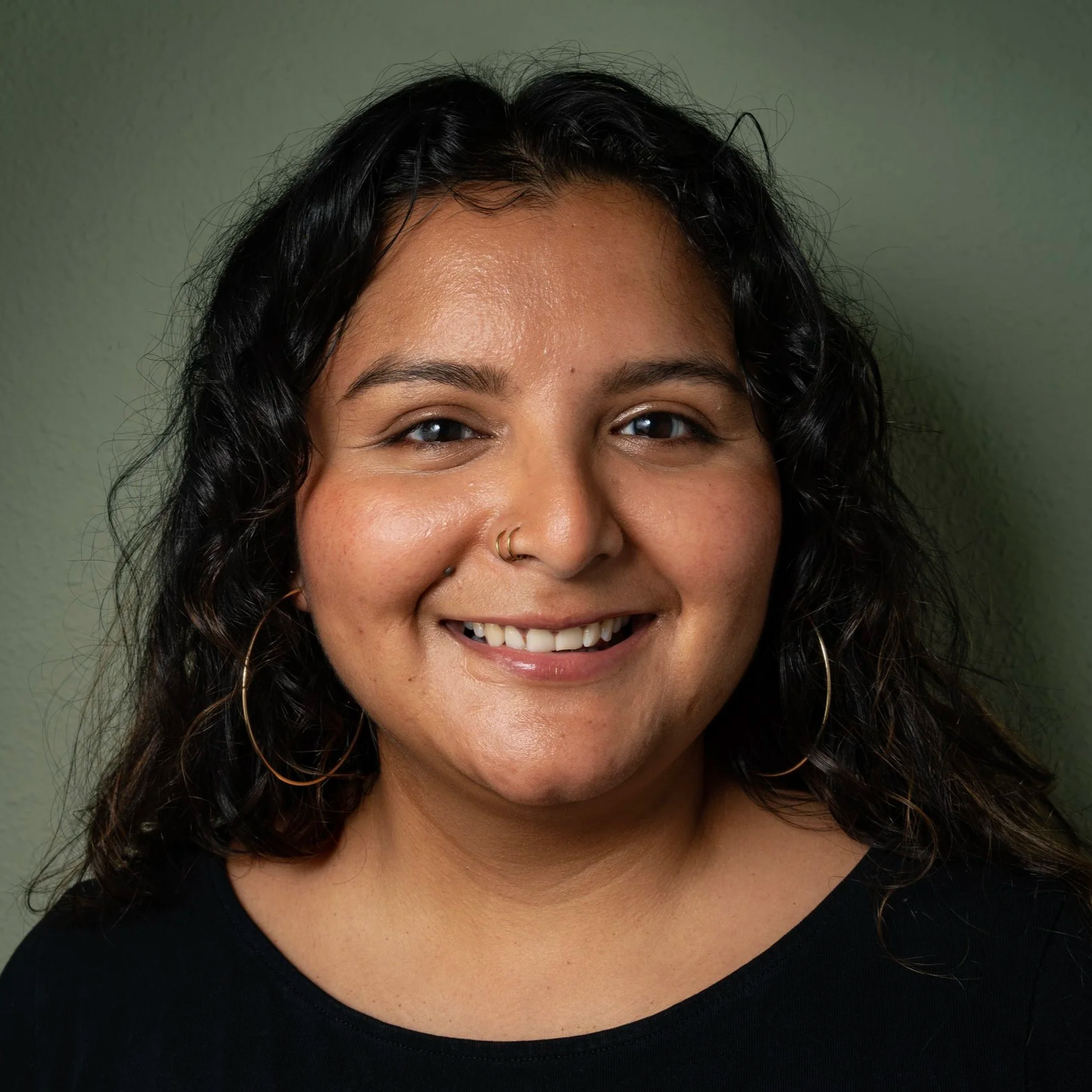 Johana Vicente, Senior Strategist, Lopez-Wagner Strategies, Close-up of a smiling woman with dark curly hair, wearing gold hoop earrings, a black top, and a nose ring, standing against a plain green wall.