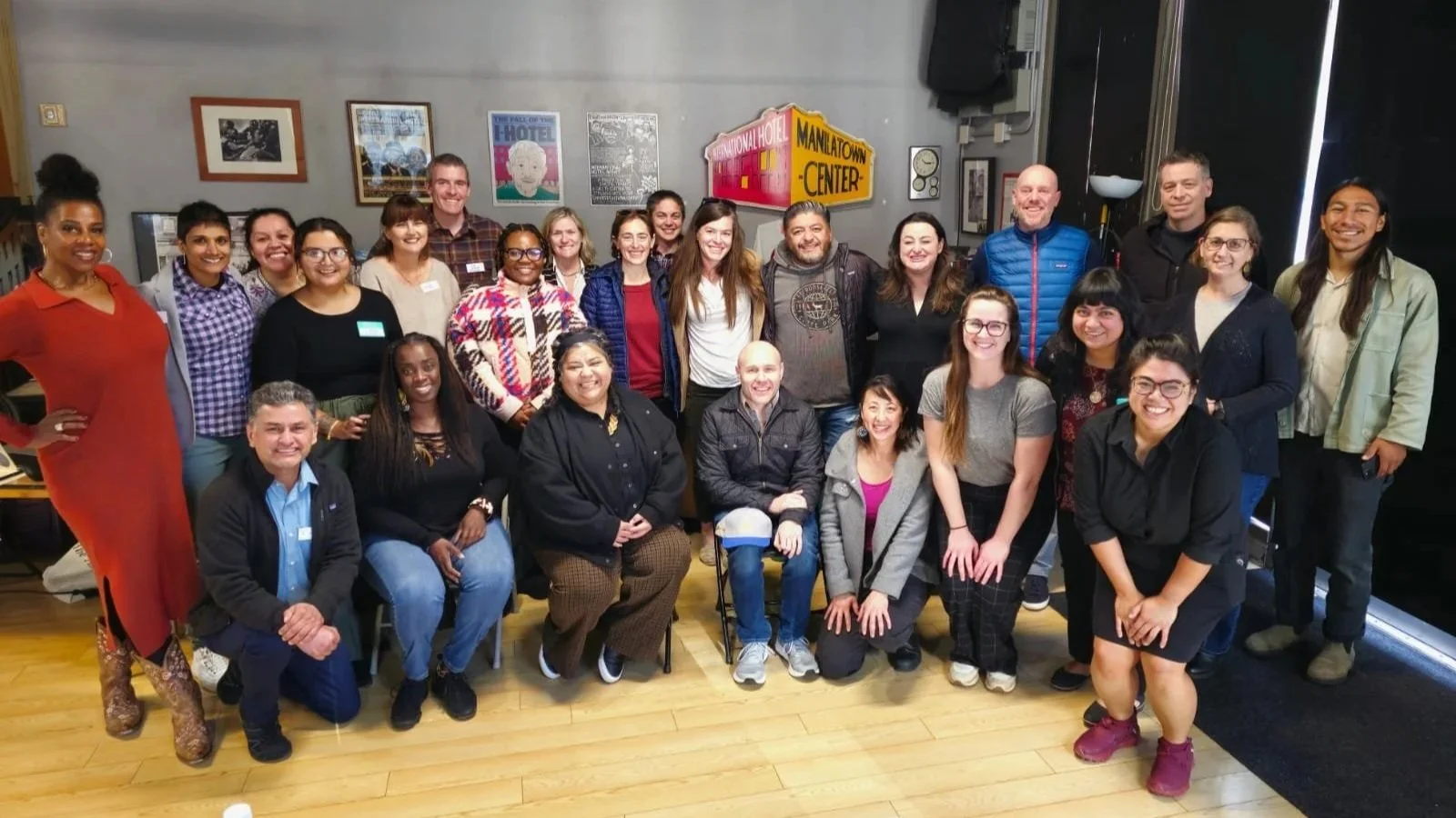Group of diverse people posing together indoors, some standing and some kneeling, with posters and signs on the wall behind them.