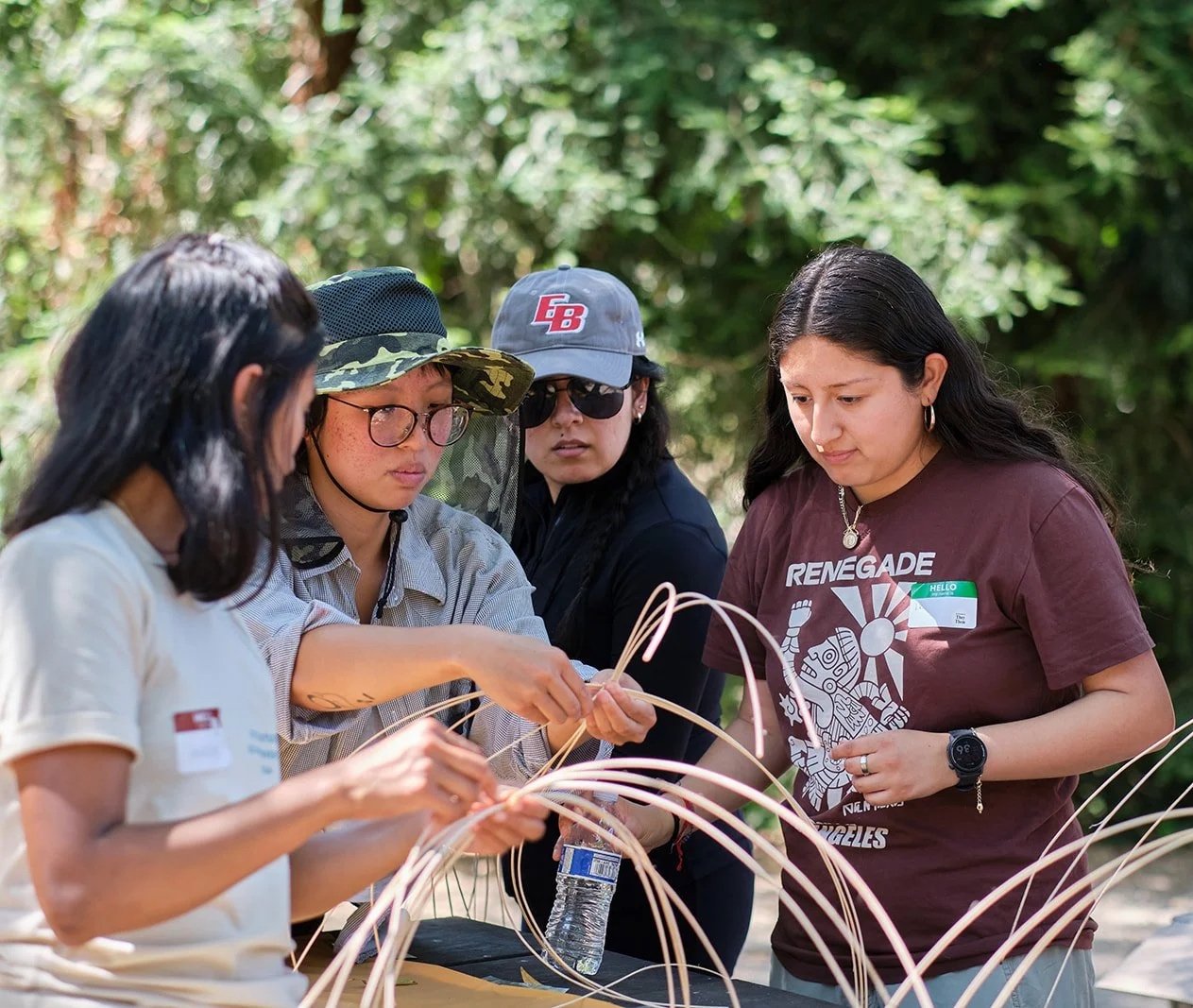 A group of four women outdoors, focused on weaving or crafting with natural material, surrounded by green foliage.