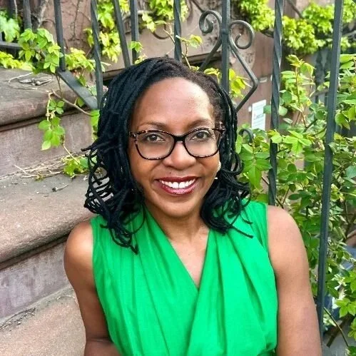 Onika Abraham Lee, Executive Director   Blue Sky Funders Forum, A woman with black curly hair and glasses wearing a green sleeveless top, smiling outdoors near a staircase with green plants and a black iron fence in the background.