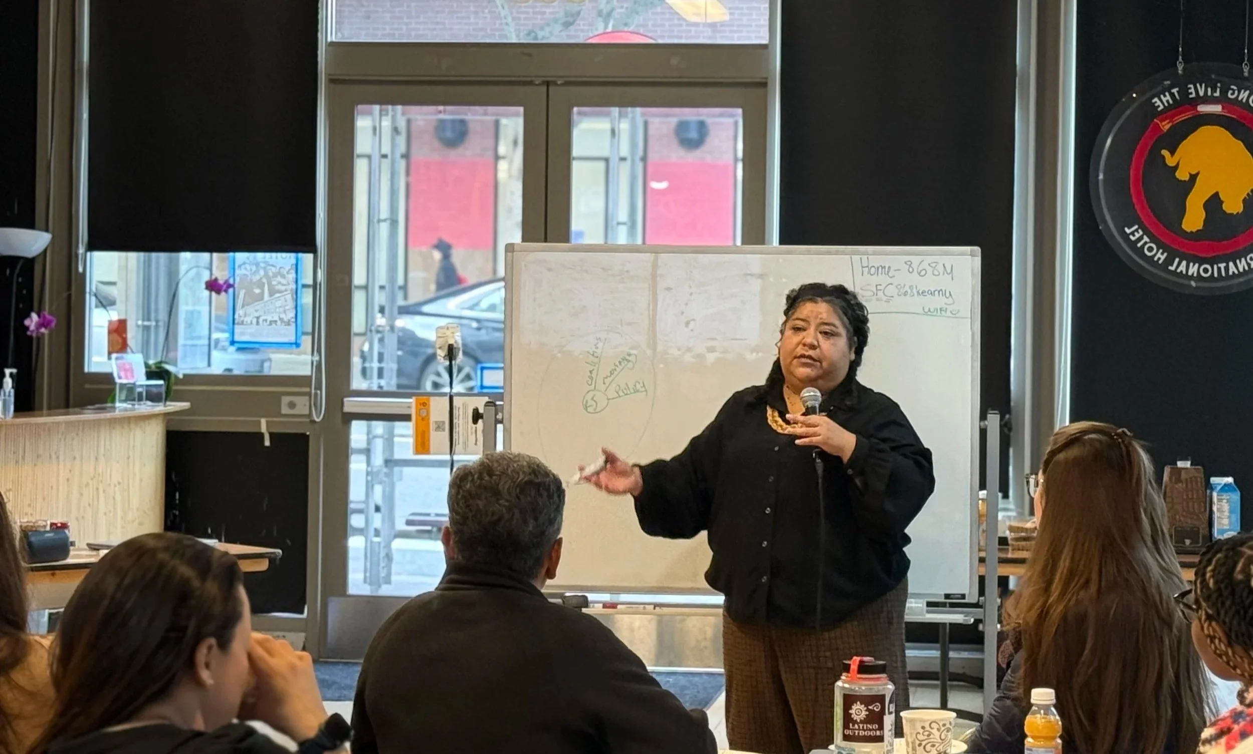 A woman giving a presentation to a group in a room with large windows. She is standing in front of a whiteboard, holding a microphone, addressing seated adults.