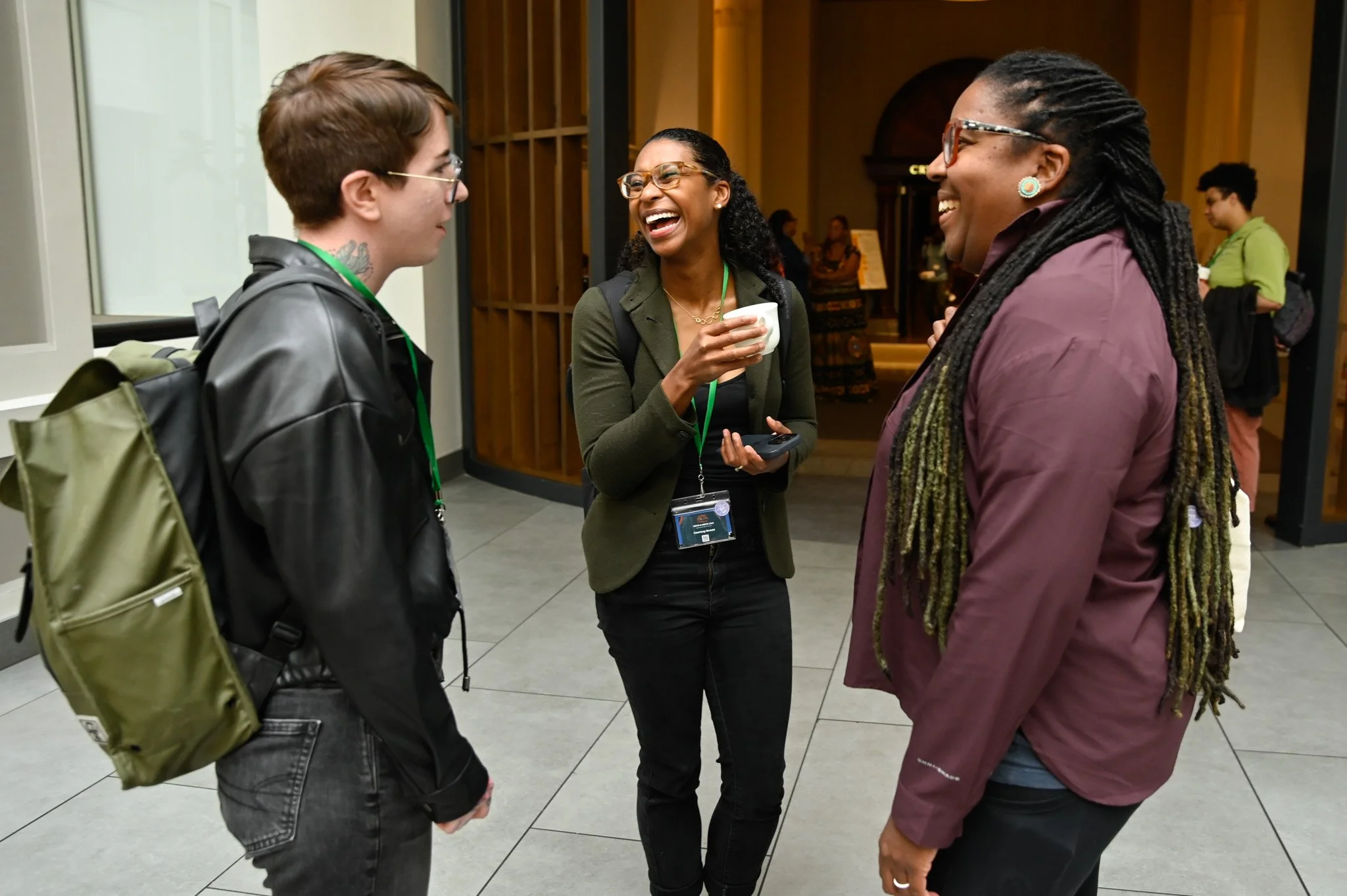 Three women talking and laughing in a public indoor space, with conference badges around their necks, and others in the background.