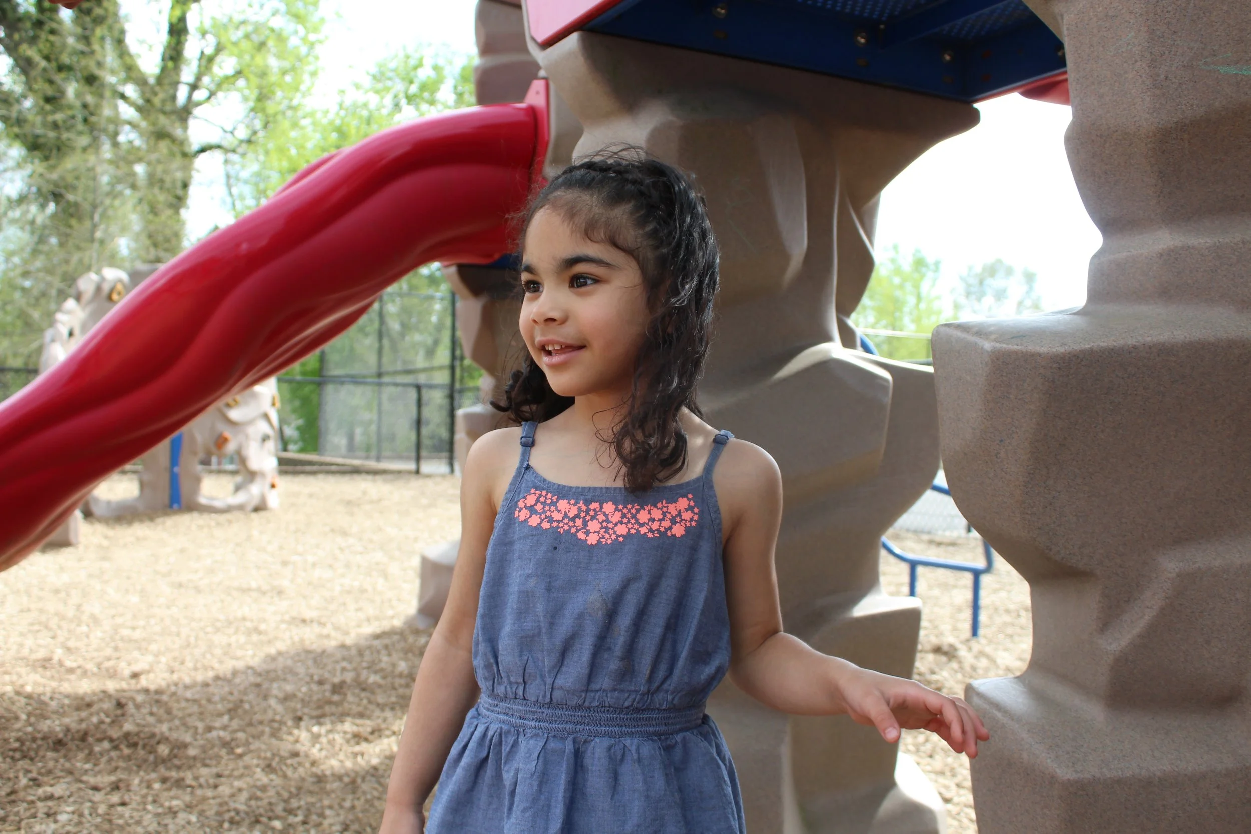 A young girl with curly dark hair standing near a playground structure, smiling and looking to her left.