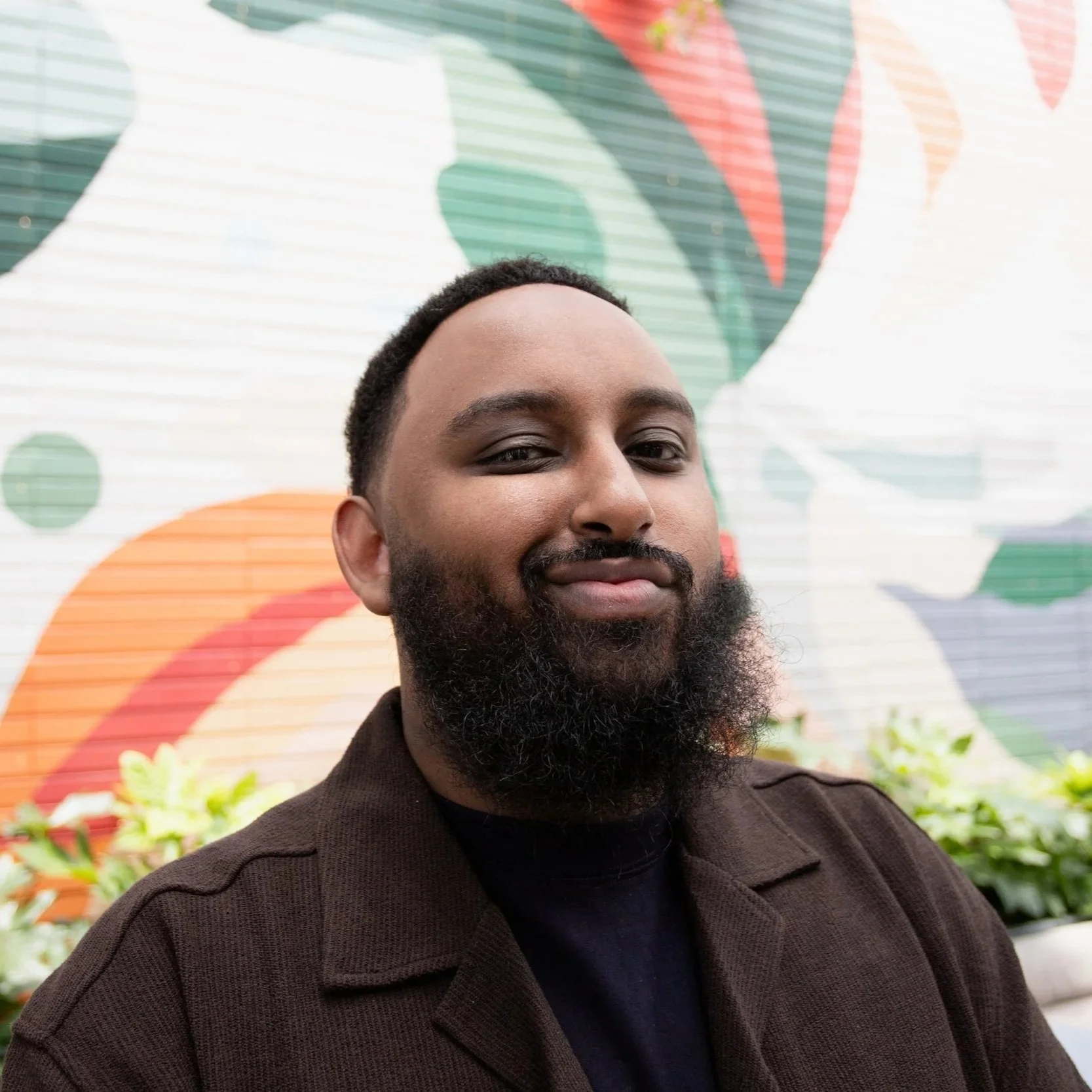 Adam Hassan, National Advocacy Manager, Close-up of a man with short curly hair and a beard, wearing a black shirt and brown coat, standing in front of a colorful mural with leaves and abstract shapes.
