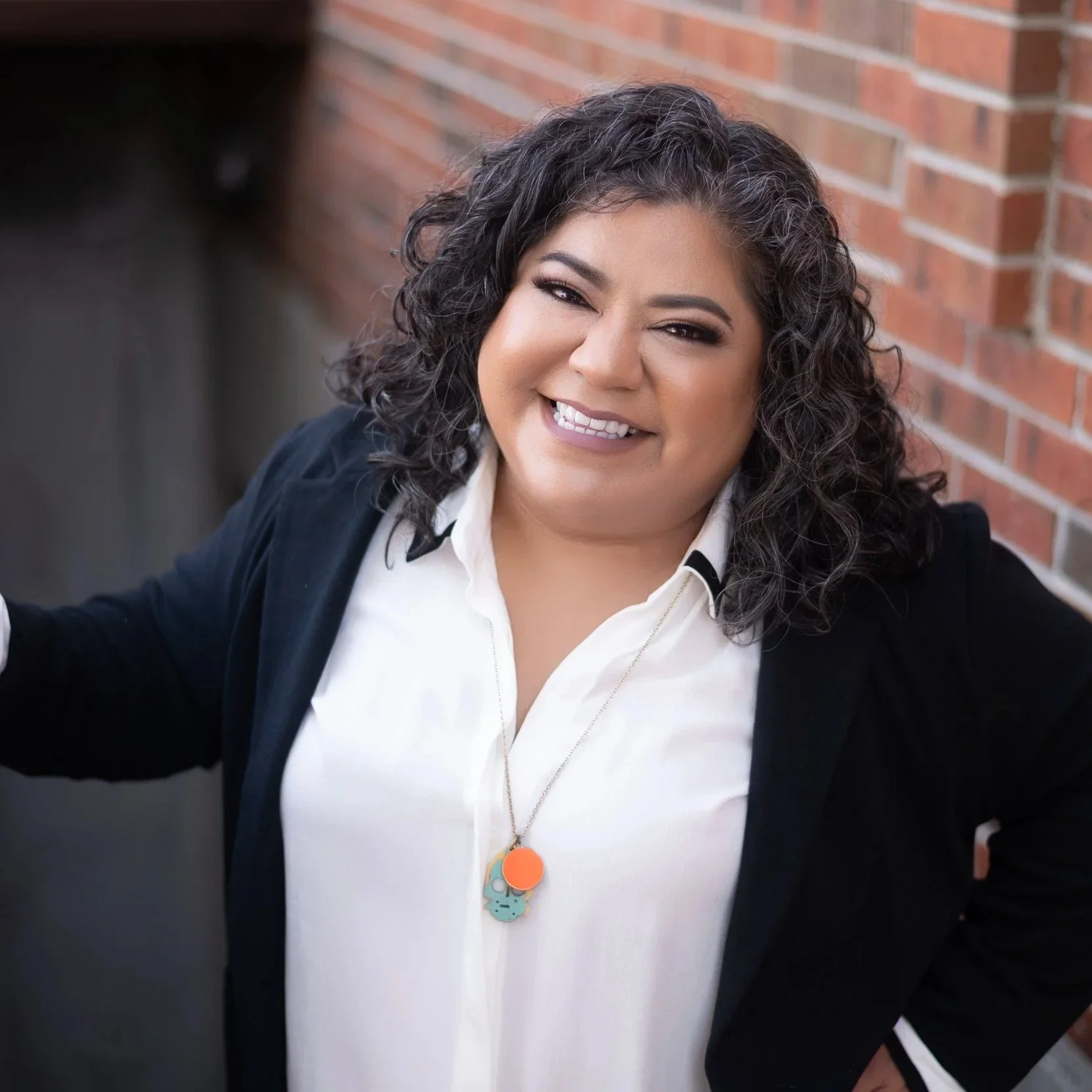 Rena Payan, Chief Program Officer, A woman with curly dark hair winks and smiles at the camera, wearing a white shirt, black blazer, and a necklace with colorful pendant, standing against a brick wall.