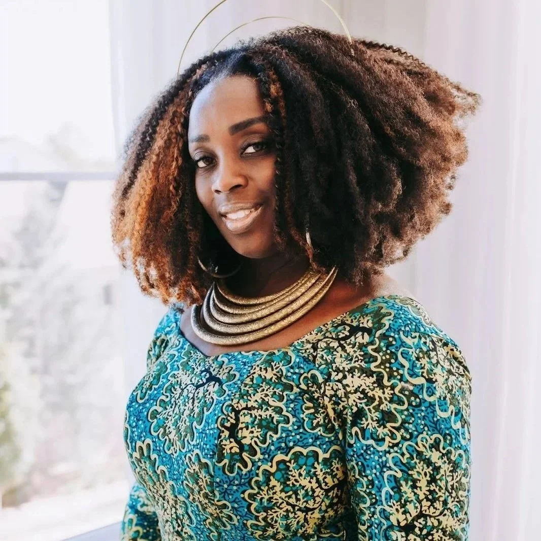 Wande Okunoren-Meadows, Executive Director  Hand, Heart, + Soul Project, A woman with curly, two-toned hair wearing a colorful, patterned dress and multiple gold necklaces, standing indoors near a window.