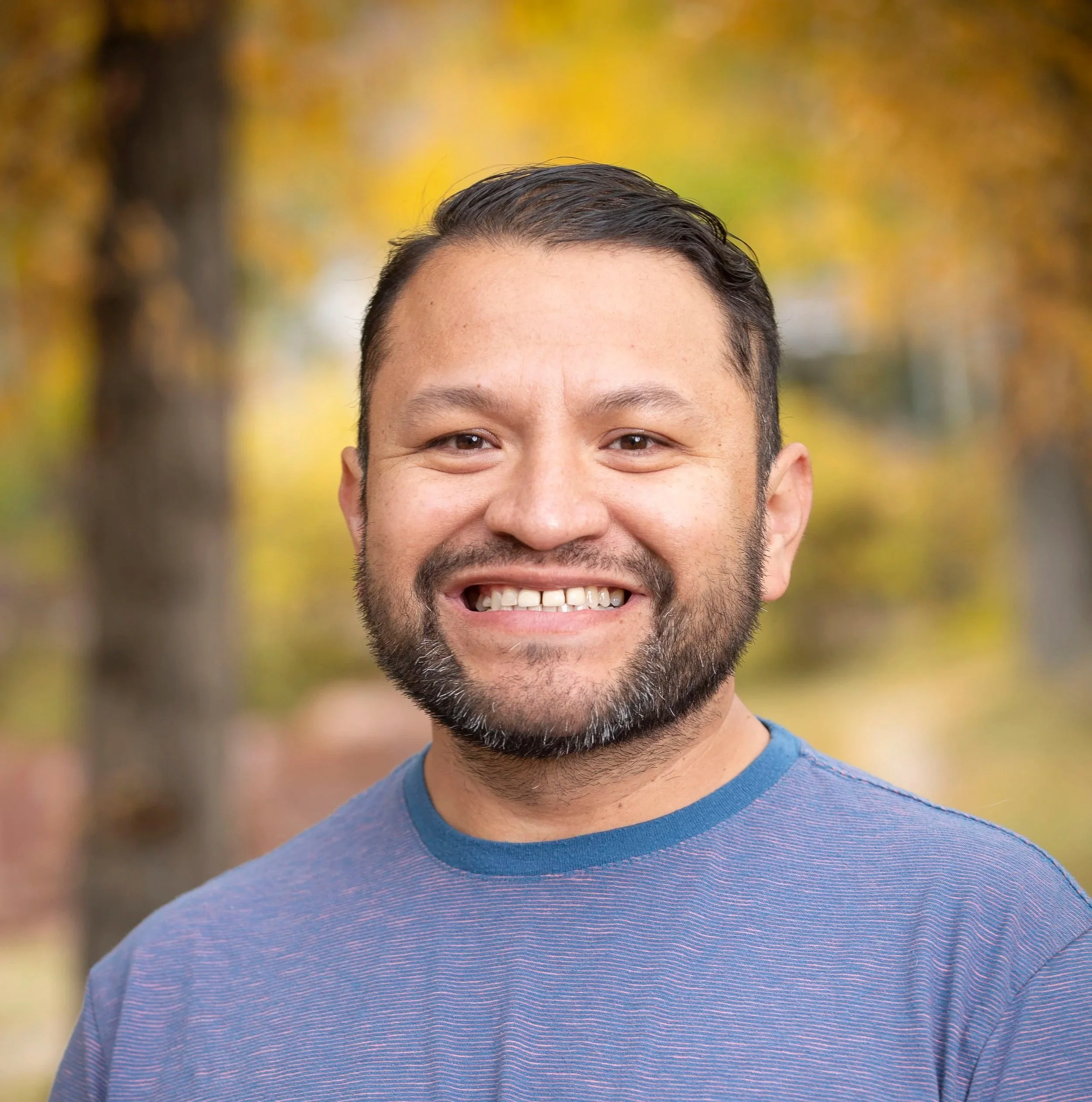 Juan D. Martinez Pineda, Director  Fresh Tracks - Aspen Institute, Close-up of a smiling man with dark hair and facial hair, wearing a purple shirt, with blurred autumn trees in the background.