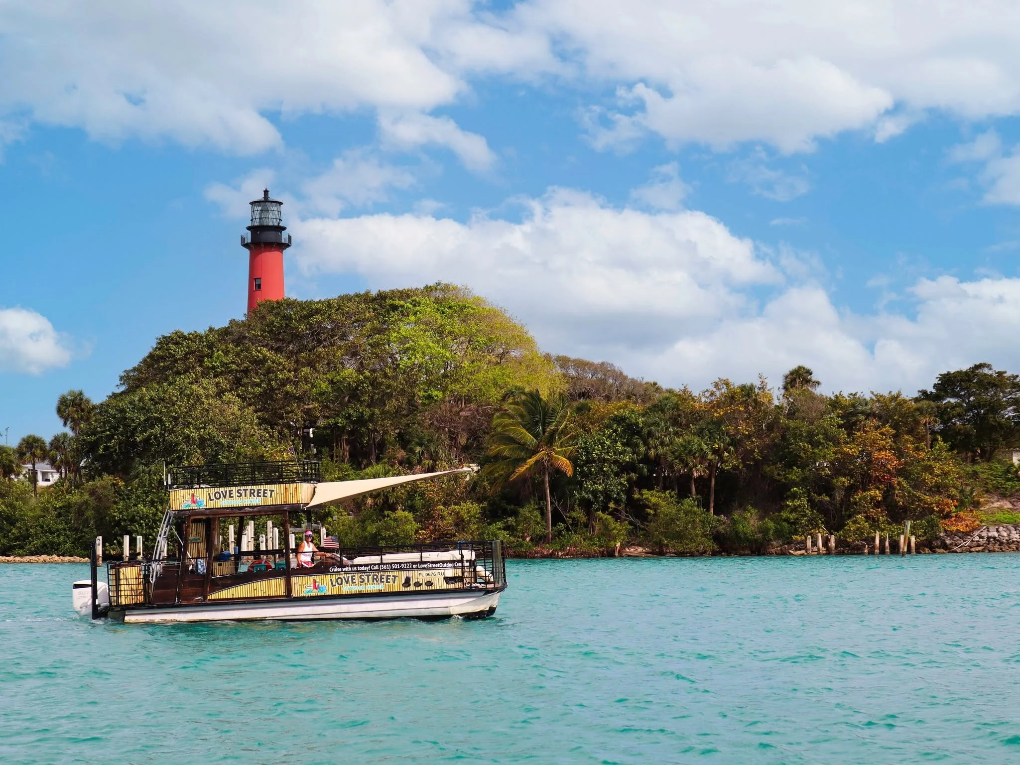 A boat with people on board cruising on turquoise water, with lush green trees and a red lighthouse on a hill in the background, under a partly cloudy sky.