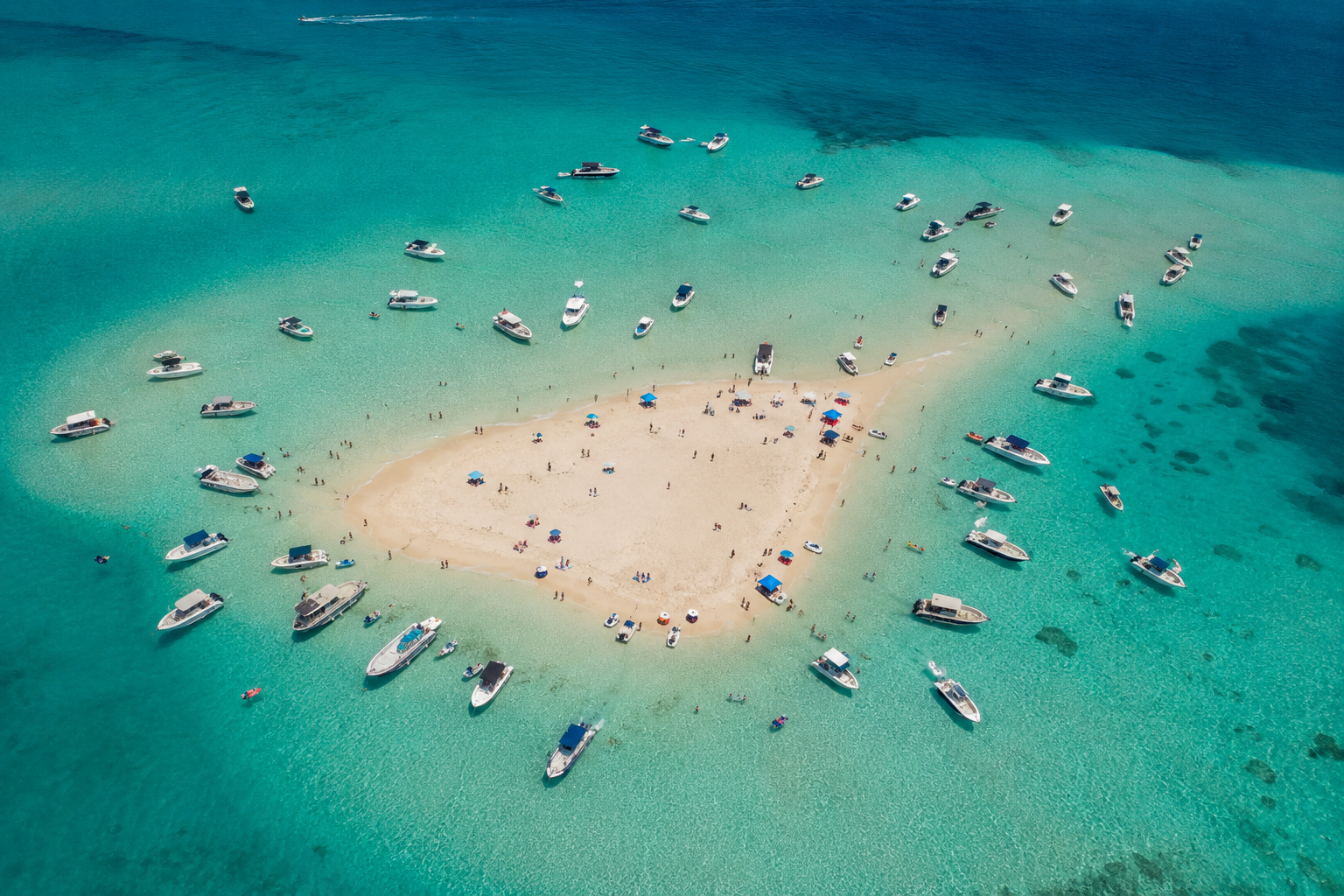 An aerial view of a sandbar in the ocean surrounded by turquoise water with numerous boats anchored around it, and people sunbathing, swimming, and relaxing on the sand.